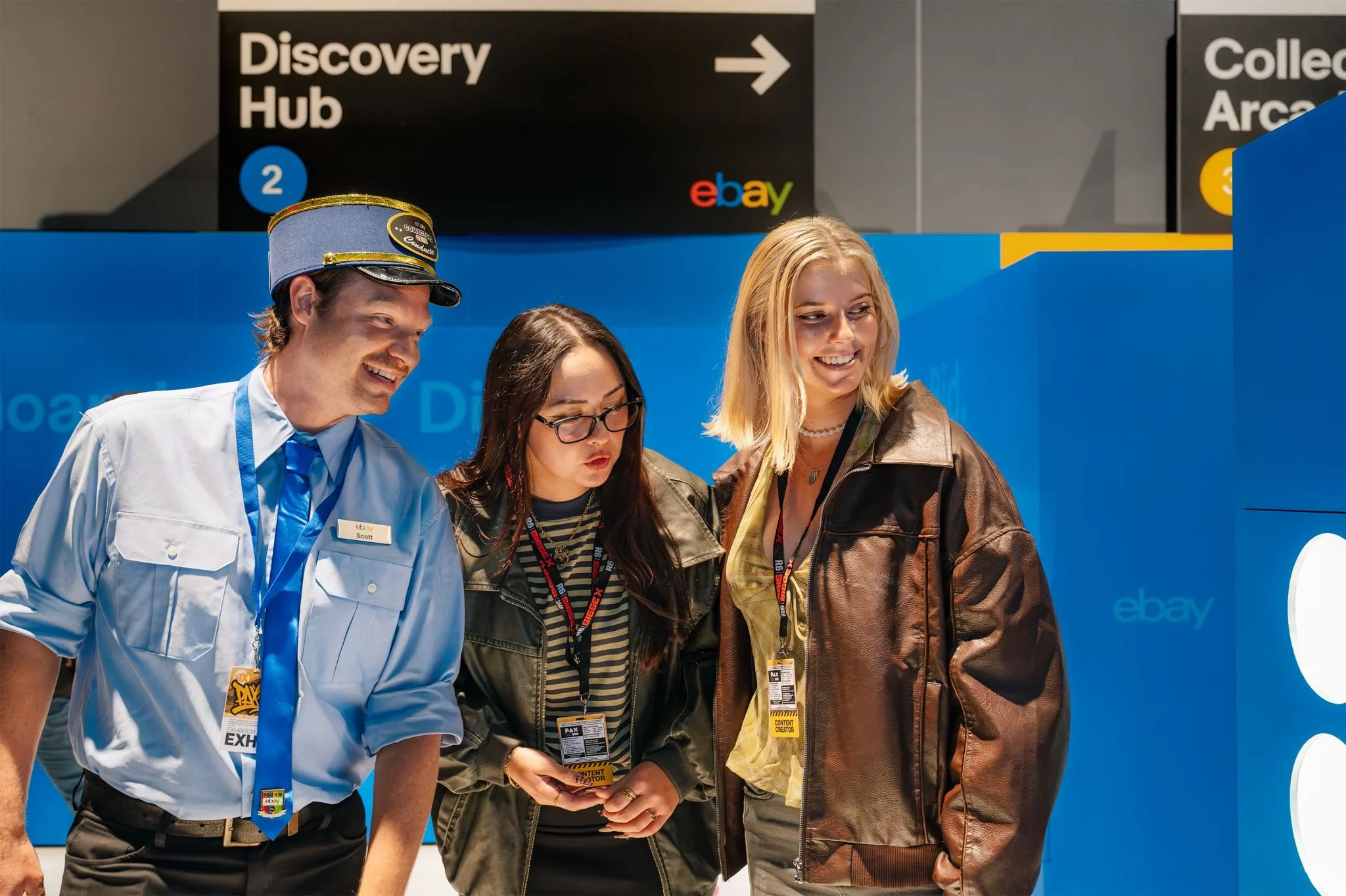 Three people, a man dressed as a train conductor, a woman wearing glasses, and a blonde woman, are standing together at an event, looking at something out of frame. There are signs behind them indicating directions to a discovery hub and eBay.
