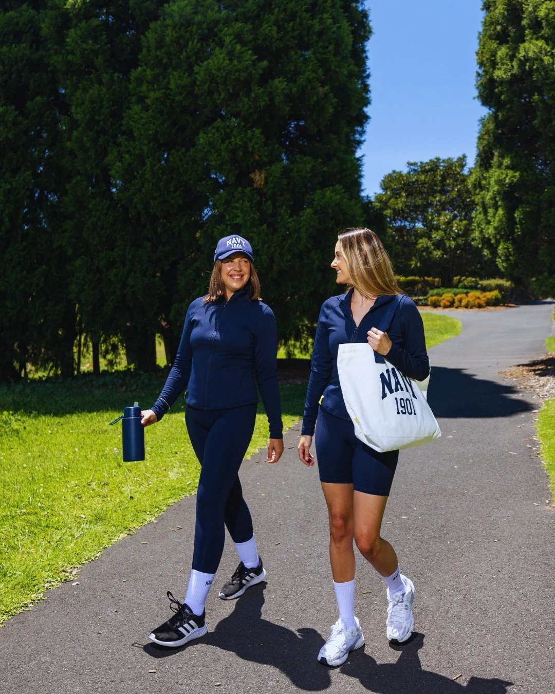 Two women in navy blue athletic wear walking on a paved trail in a park, smiling and talking. One woman is carrying a white tote bag with the word 'NAVY' and the number '190' on it, while the other holds a navy water bottle. They are surrounded by green trees and grass.