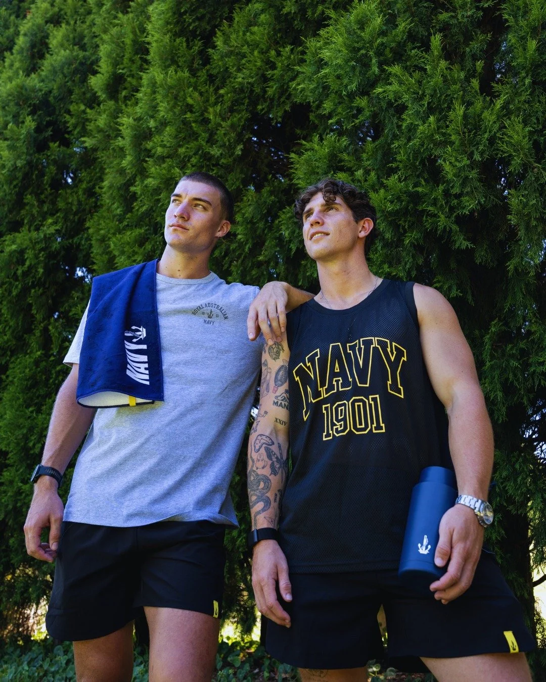 Two young men standing outdoors in front of green trees, wearing Navy-themed athletic clothing, one with tattoos and holding a water bottle, the other with a towel draped over his shoulder.