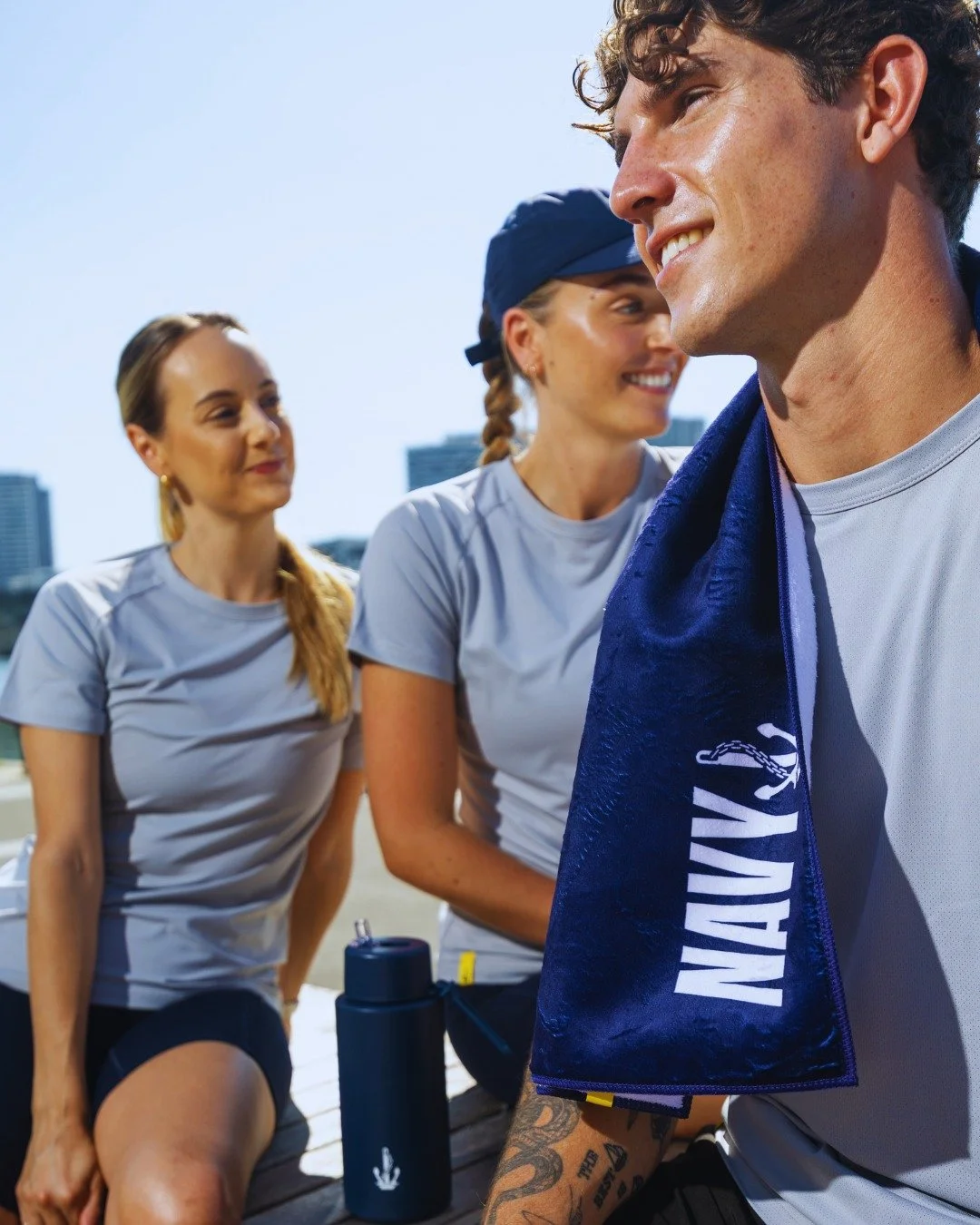 Three young adults, two women and one man, are sitting outdoors on a sunny day, dressed in athletic gear, with buildings visible in the background. The man has a towel with the word 'NAVY' draped over his shoulder, and a water bottle is in front of them.