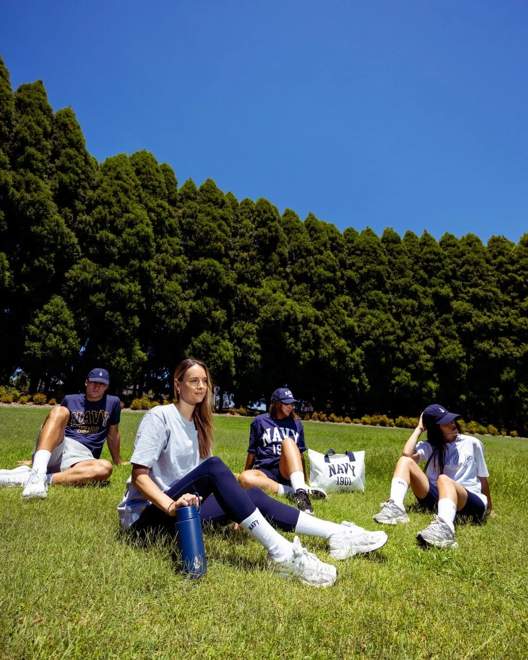 Four young women sitting on the grass in a park, wearing navy athletic shirts, shorts, caps, and sneakers, with trees and a blue sky in the background.