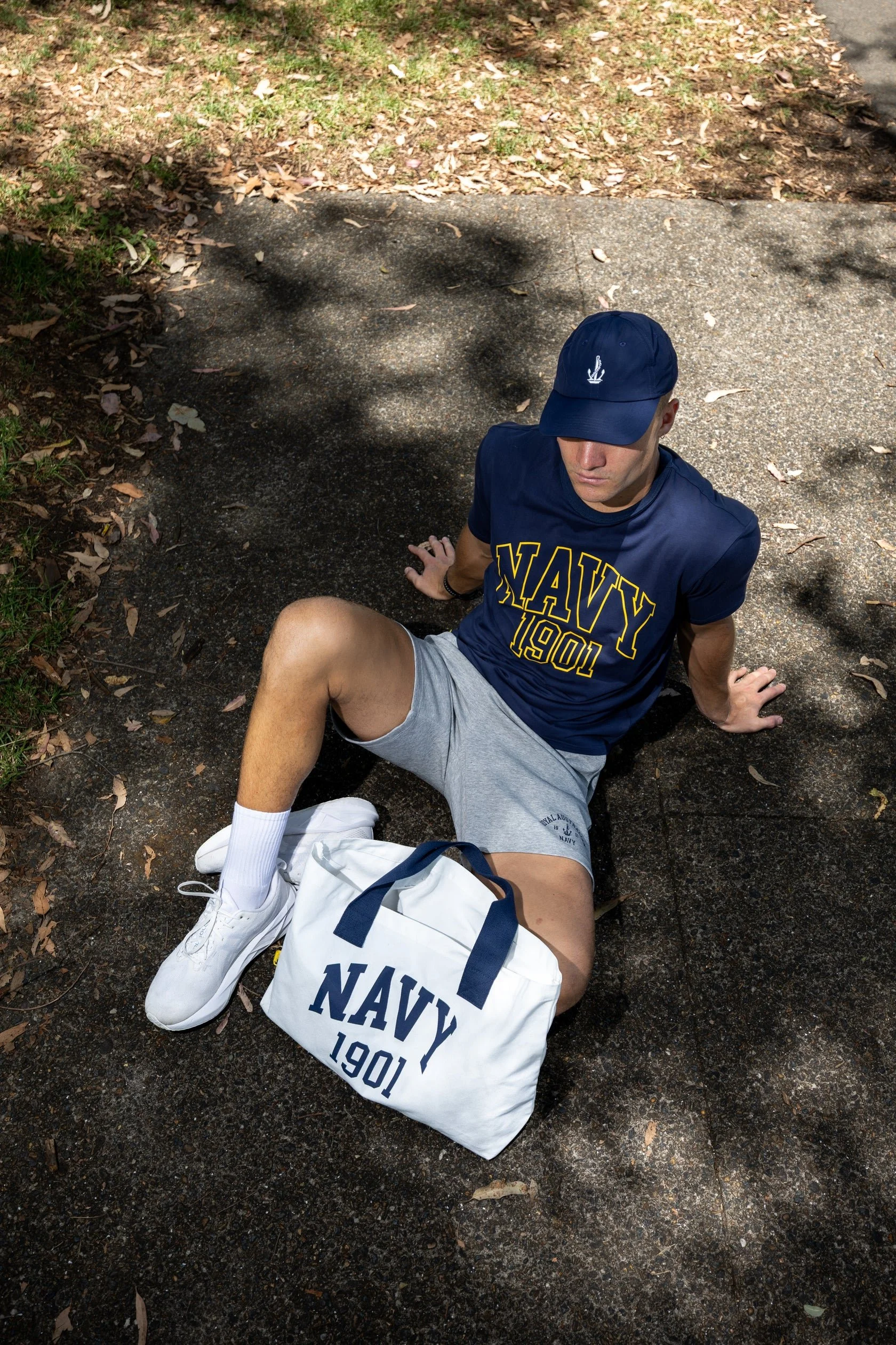 A young man is sitting on the ground outdoors, wearing a navy blue baseball cap and a navy blue T-shirt with 'NAVY 1901' written on it. He has on gray shorts, white sneakers, and appears to be resting or relaxing next to a navy blue and white bag with the same 'NAVY 1901' branding. The ground is a mix of concrete and dirt with some fallen leaves, and trees cast shadows over the scene.