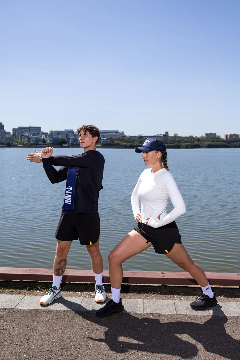 A man and a woman in athletic clothing stretching near a body of water with a city skyline in the background. The man is pulling his arm across his chest, and the woman is doing a wide-leg stretch with her hands on her hips. Both are wearing navy hats and matching athletic gear with 'NAVY' written on their accessories.