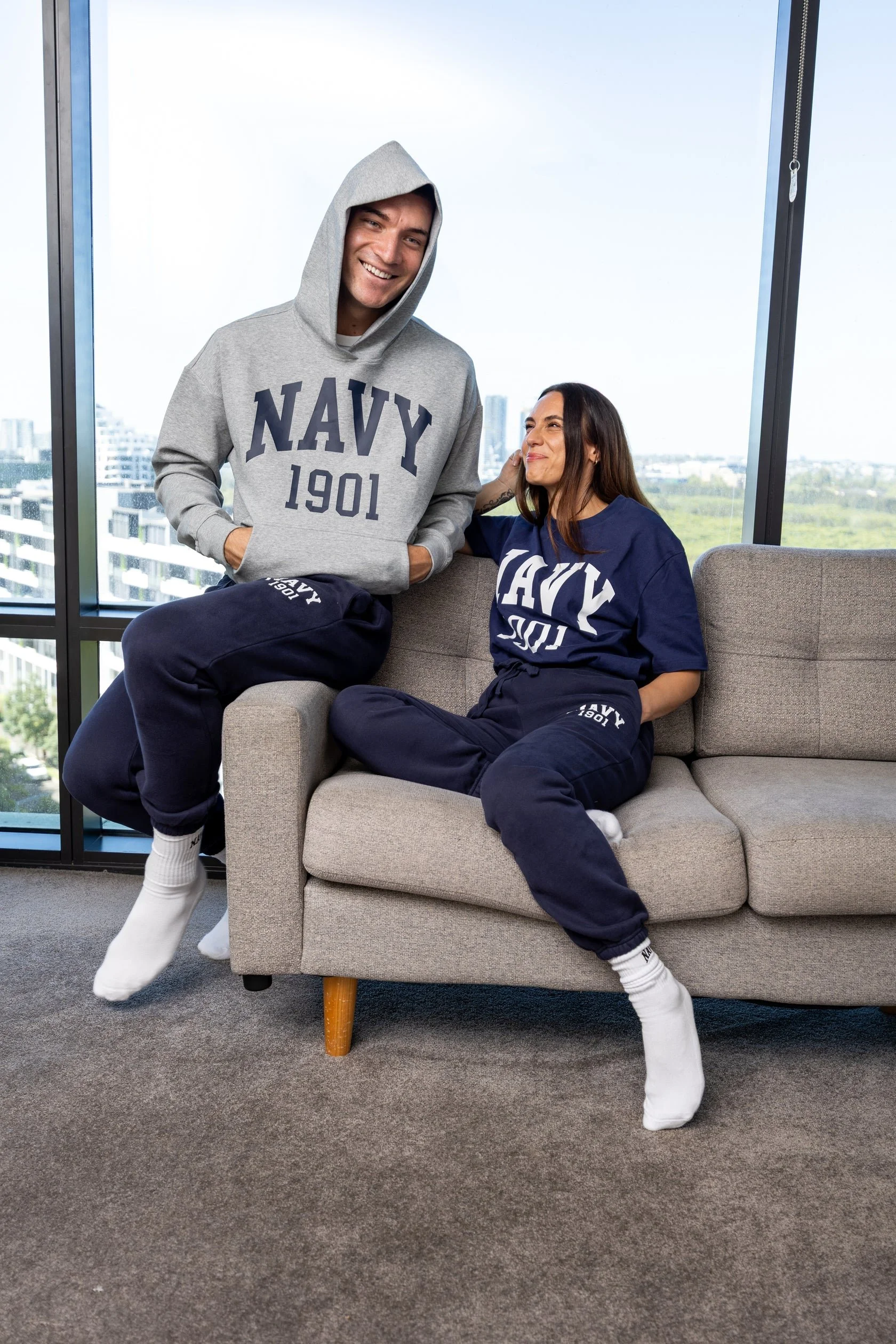 A man standing and a woman sitting on a sofa inside a modern office with large windows showing a city skyline. They are wearing navy-themed sweatshirts and sweatpants.
