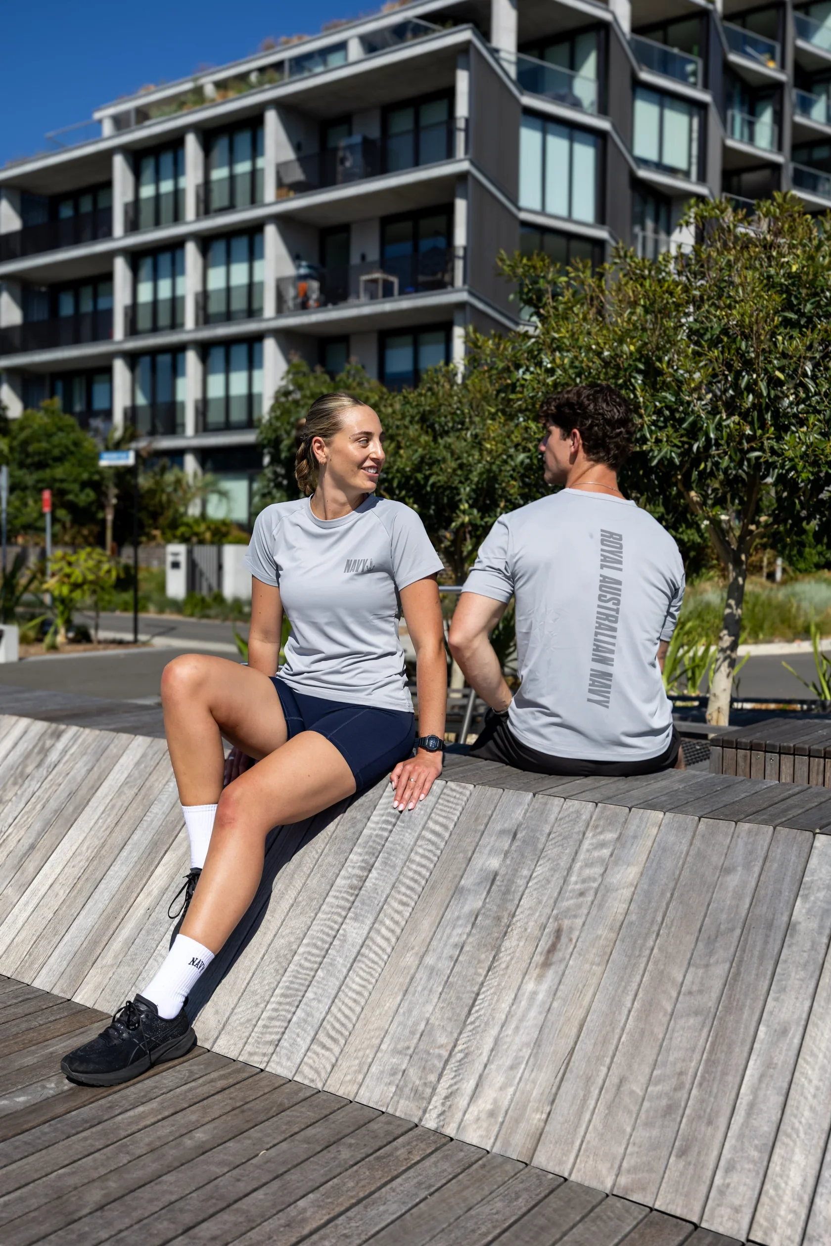 Two people, a woman and a man, are sitting on a curved wooden bench outdoors. They are wearing athletic clothing with the navy and Royal Australian Navy logos. The woman is smiling and looking at the man, who is facing away from the camera. There are trees, bushes, and a modern apartment building in the background under a clear blue sky.