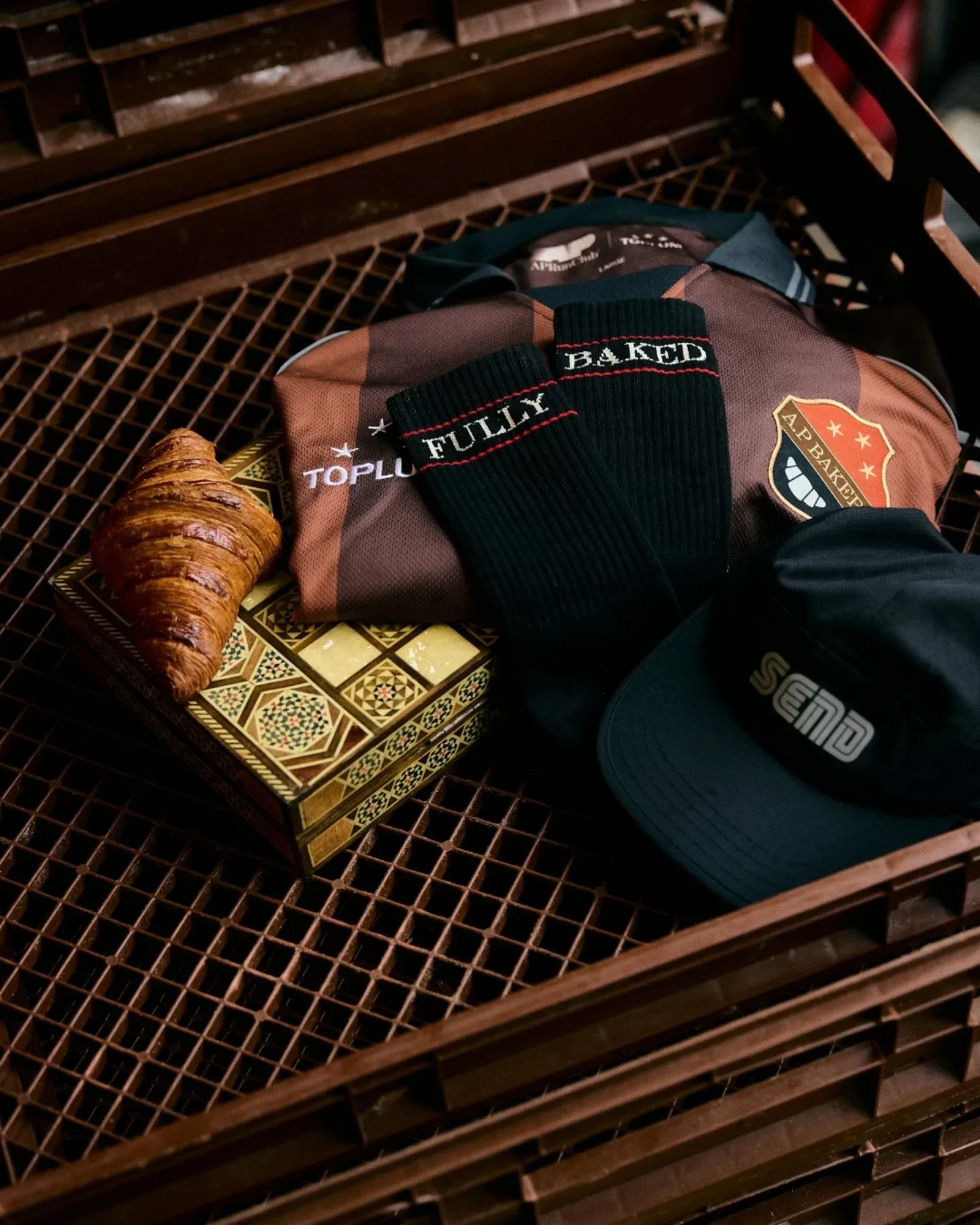 A croissant, a box with geometric patterns, a brown and orange sports bag with a shield logo, a black knit cap, and a pair of black socks with red and white text, all on a metal grate table.