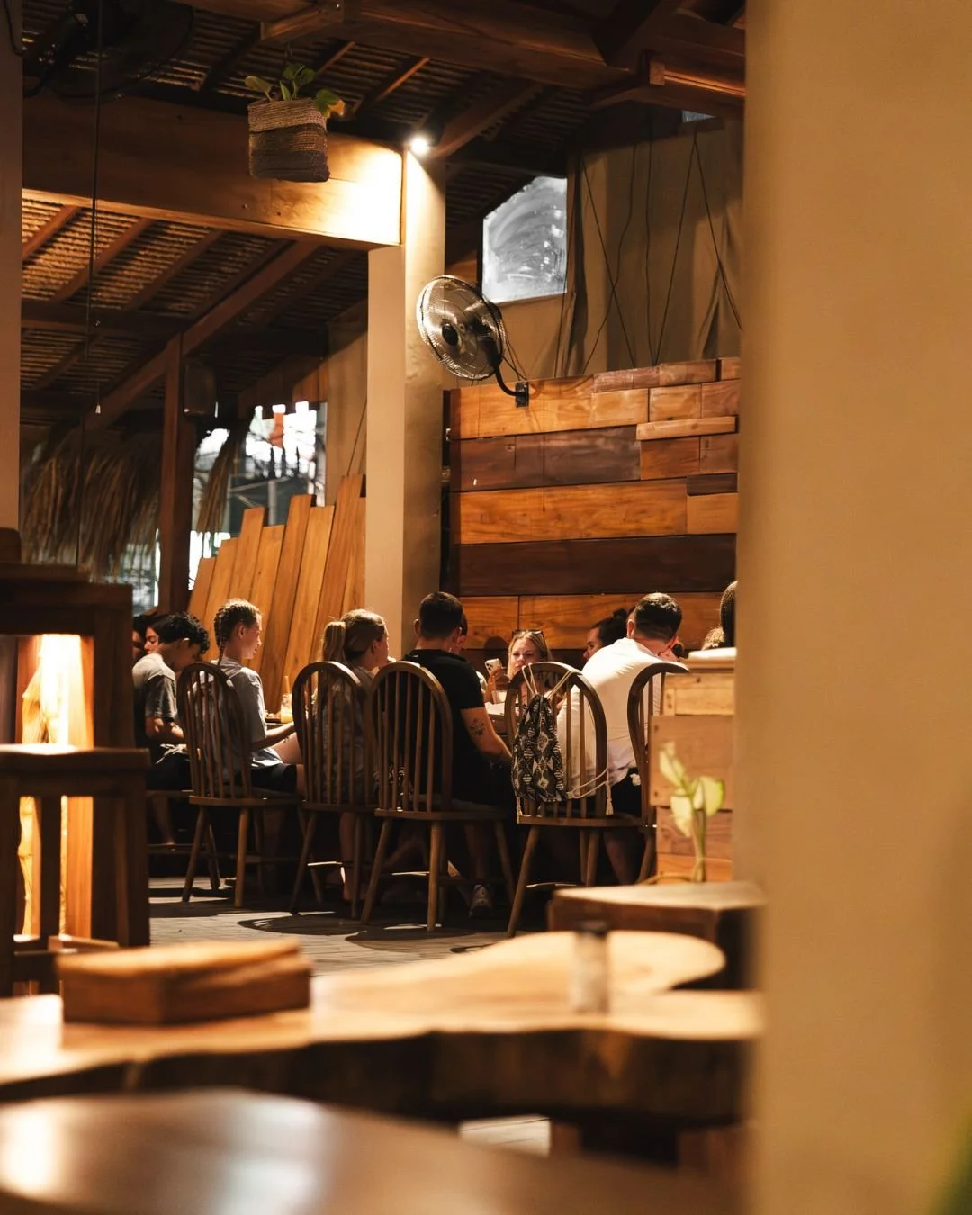 Group of people dining at a restaurant with wooden interior decor, warm lighting, and a large fan on the wall.