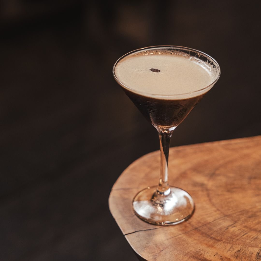A black coffee cocktail topped with a coffee bean served in a martini glass on a wooden surface.