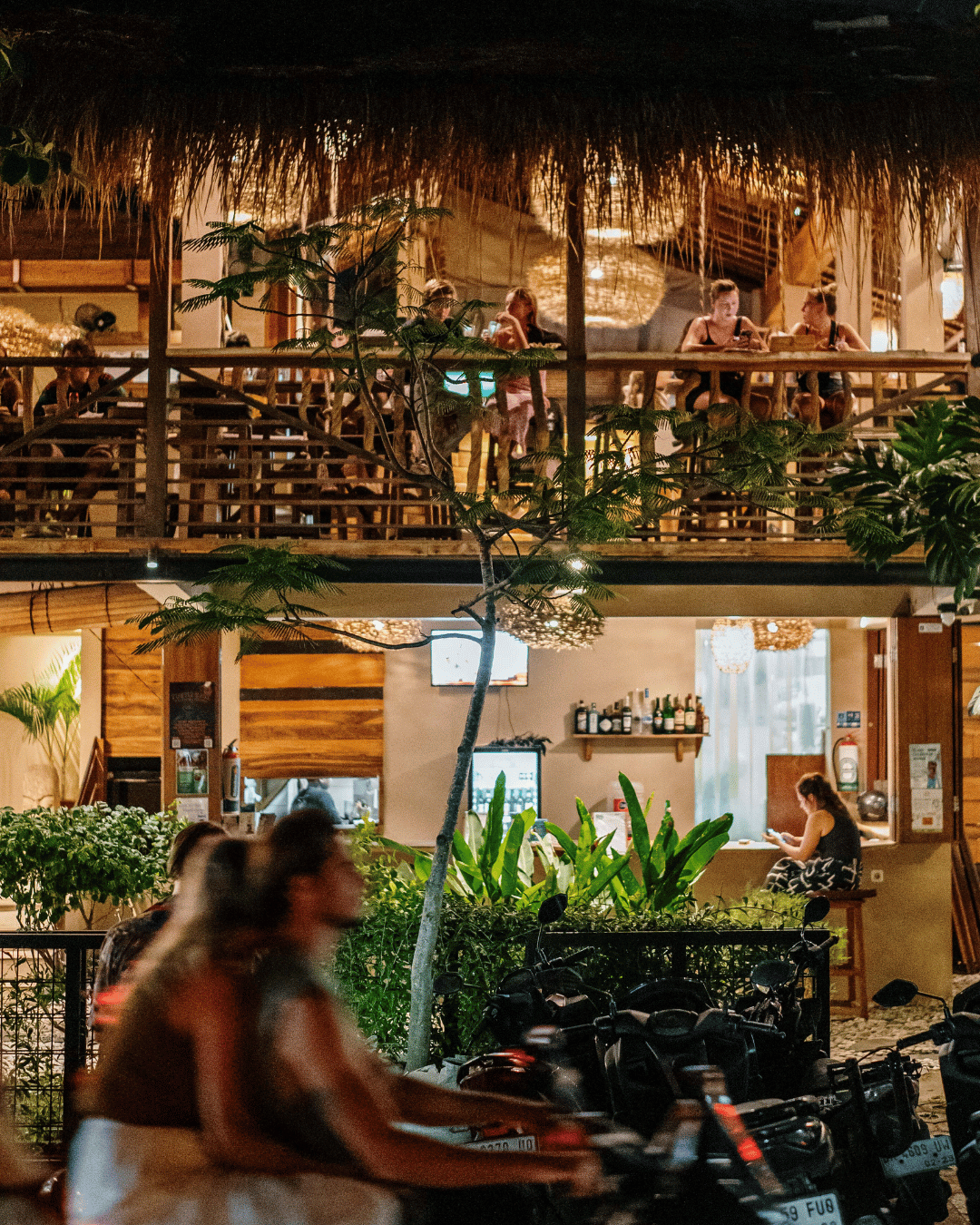Night scene of a Treehouse's two-story restaurant and bar with wooden decor and thatched roof, people sitting on the upper balcony and lower seating area, some blurred pedestrians and motorcycles in foreground, indoor plants and warm lighting.