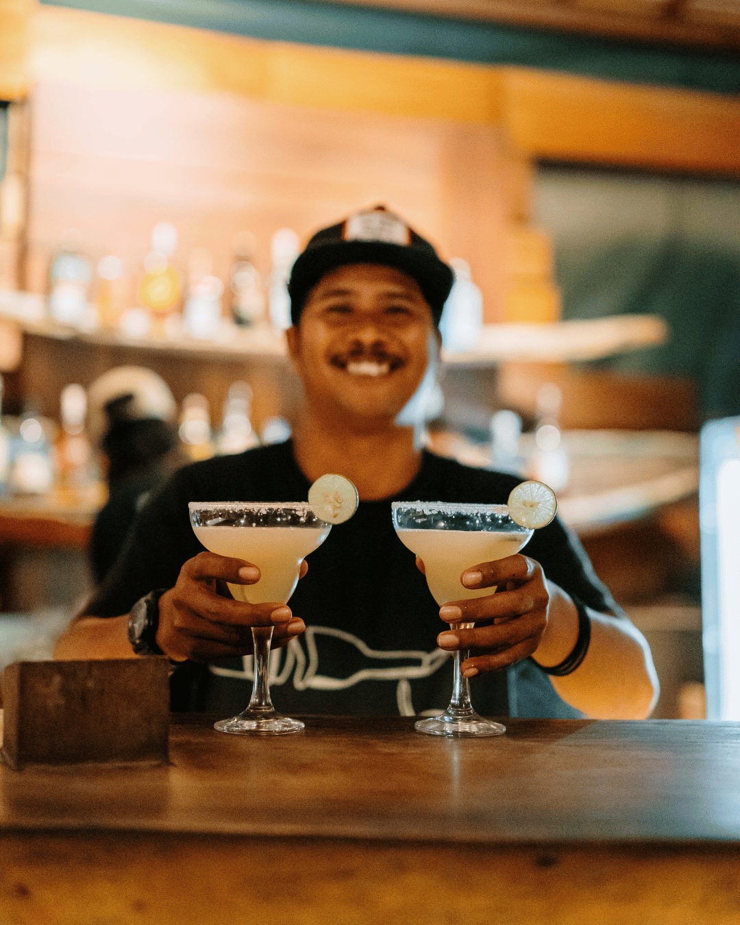A Treehouse bartender with a mustache and a baseball cap holding two cocktails with lemon slices in a bar or restaurant.