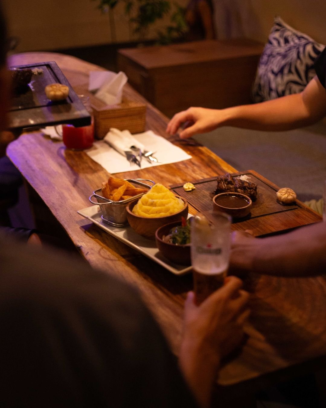 A restaurant table set with various dishes including mashed potatoes, fried food, a glass of red drink, a glass of beer, and a small bowl of sauce. There are also utensils, napkins, and a tissue box on the table, with people seated around.