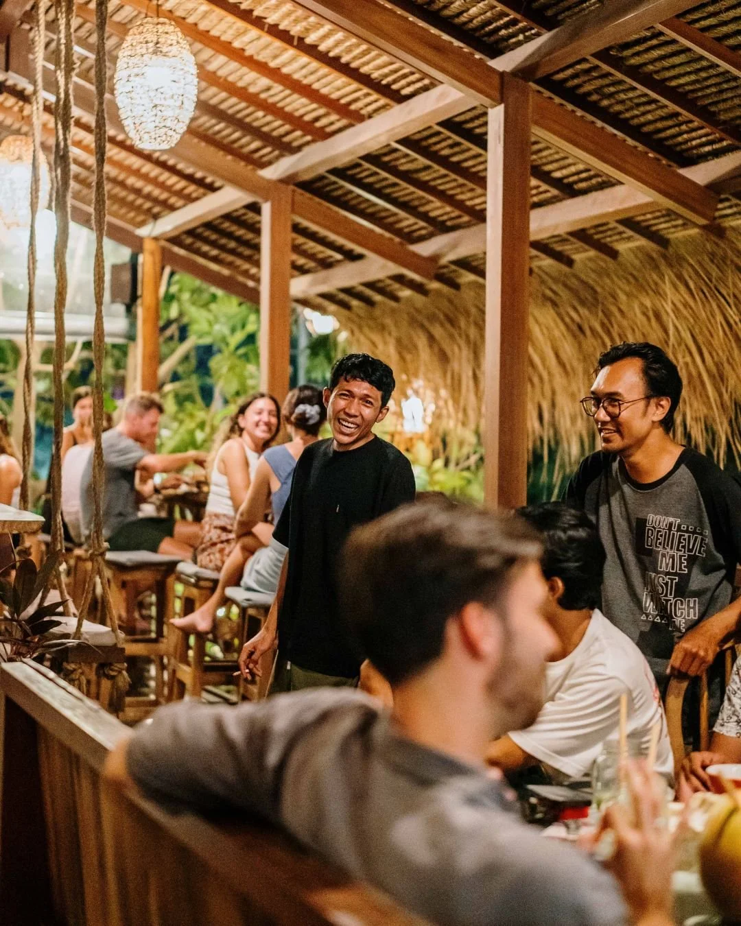 People enjoying a social gathering in a tropical-themed restaurant or bar, with wooden furniture and thatched roofing.