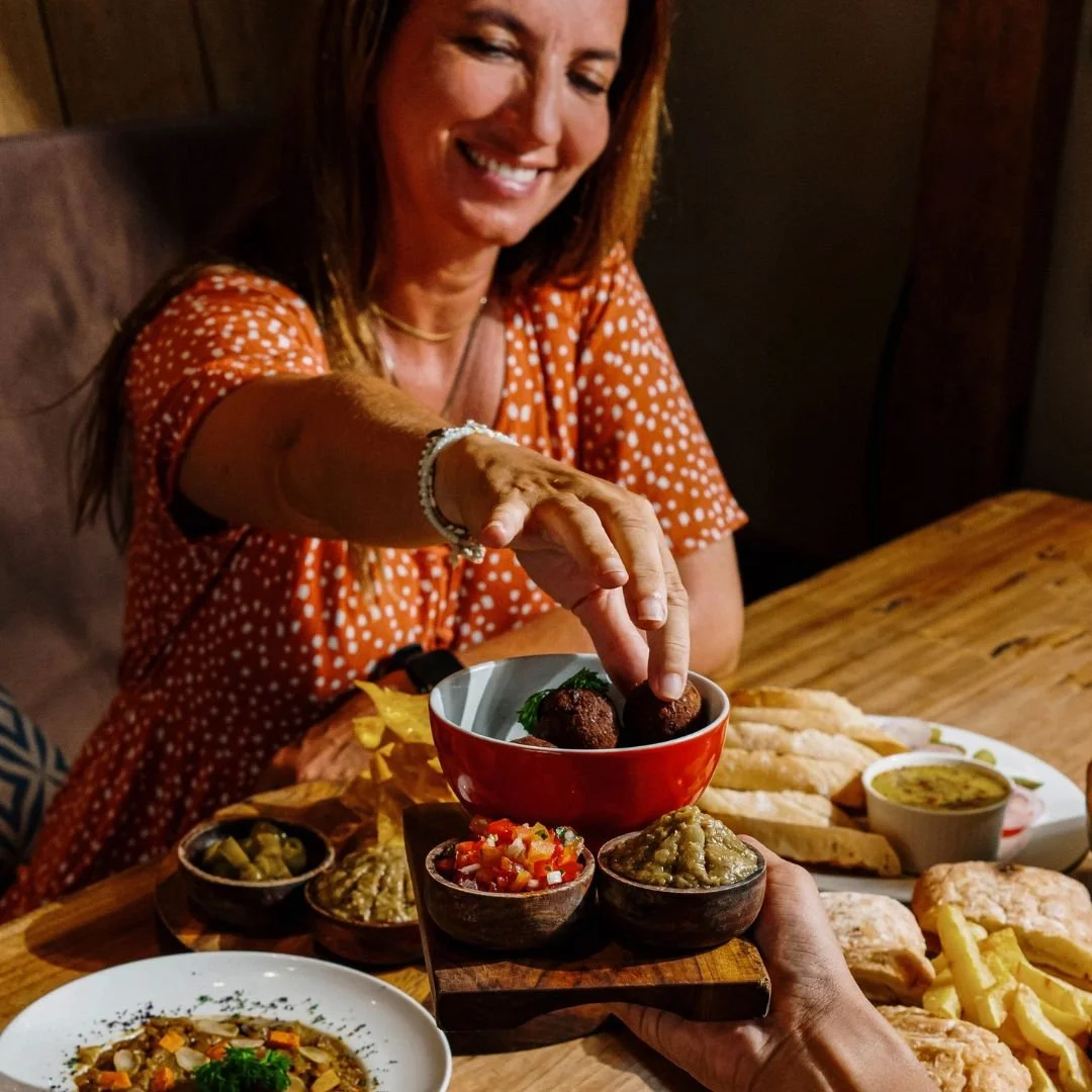 A woman in an orange polka-dot dress is smiling and reaching for food at a table with various dishes, including a bowl of falafel, dipping sauces, sliced bread, and a bowl of soup.