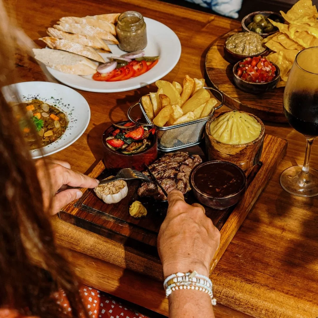 A person serving themselves grilled meat and dipping sauce from a wooden platter with various Mexican dishes and drinks on a wooden table.