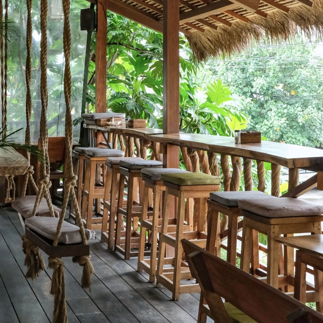 A wooden outdoor balcony with bar-height stools with cushions, a swing with a cushion hanging from ropes, and lush green plants in the background.