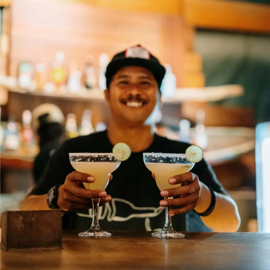 A smiling man in a baseball cap holding two cocktails garnished with lime slices in a bar setting.