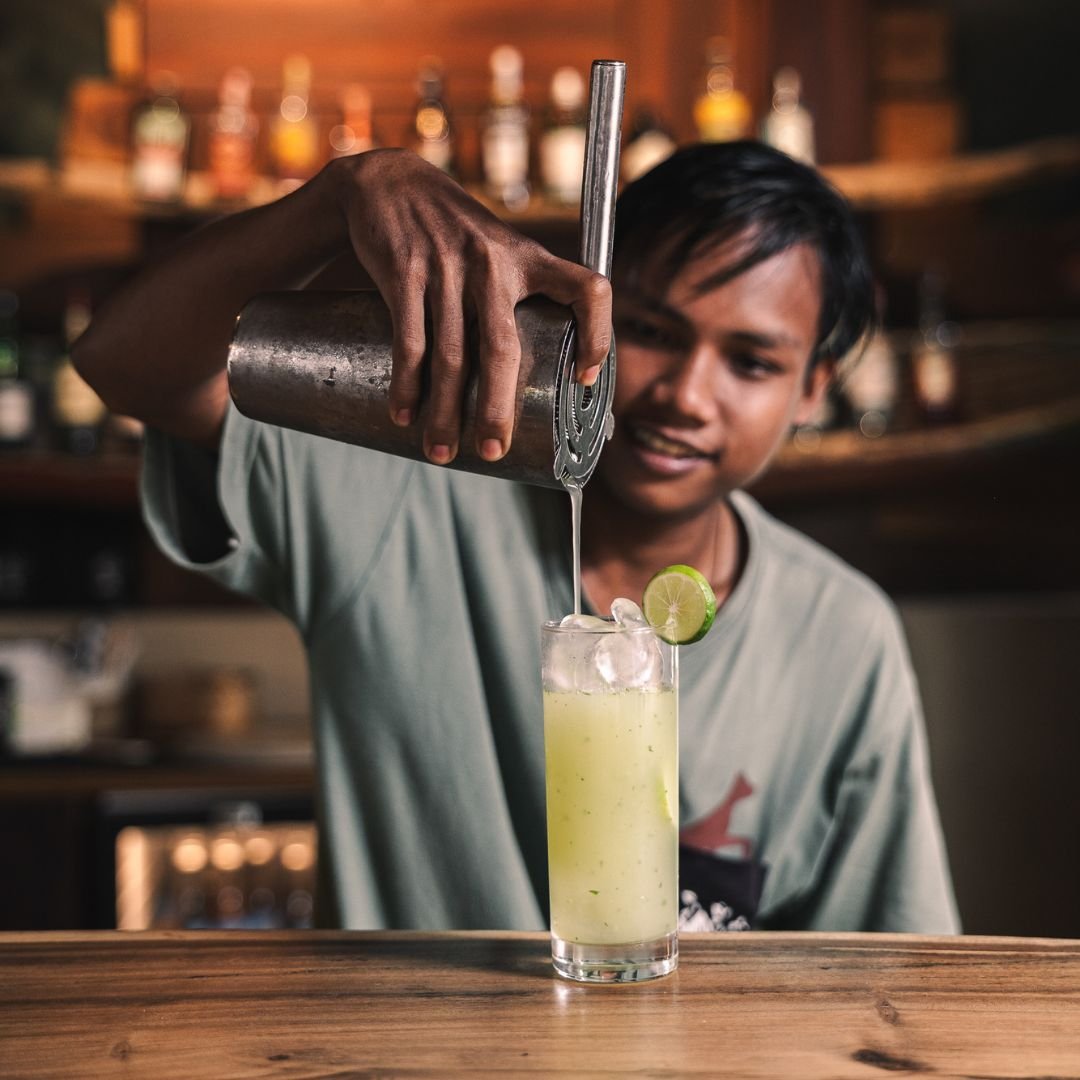Bartender pouring a yellow cocktail into a glass garnished with a lime slice in a bar setting.
