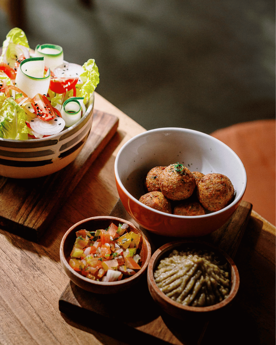 Assorted dishes including a salad with lettuce, and cucumber ribbons, a bowl of chickpea falafel balls, a small bowl of chopped tomato salsa, and another bowl of guacamole, all arranged on a wooden serving tray at Treehouse Restaurant in Lombok