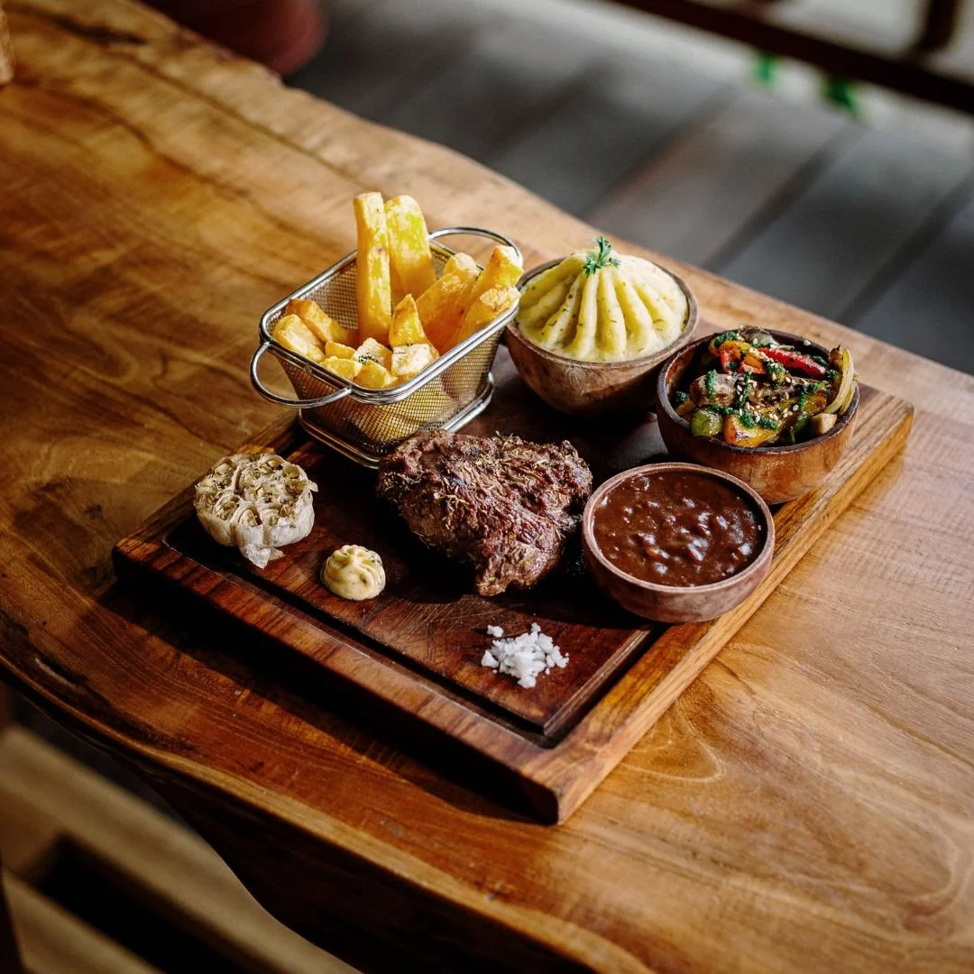 A wooden platter with French fries in a small basket, mashed potatoes, sautéed vegetables, a grilled steak, a bowl of gravy, garlic, and a dollop of mustard on a wooden table.