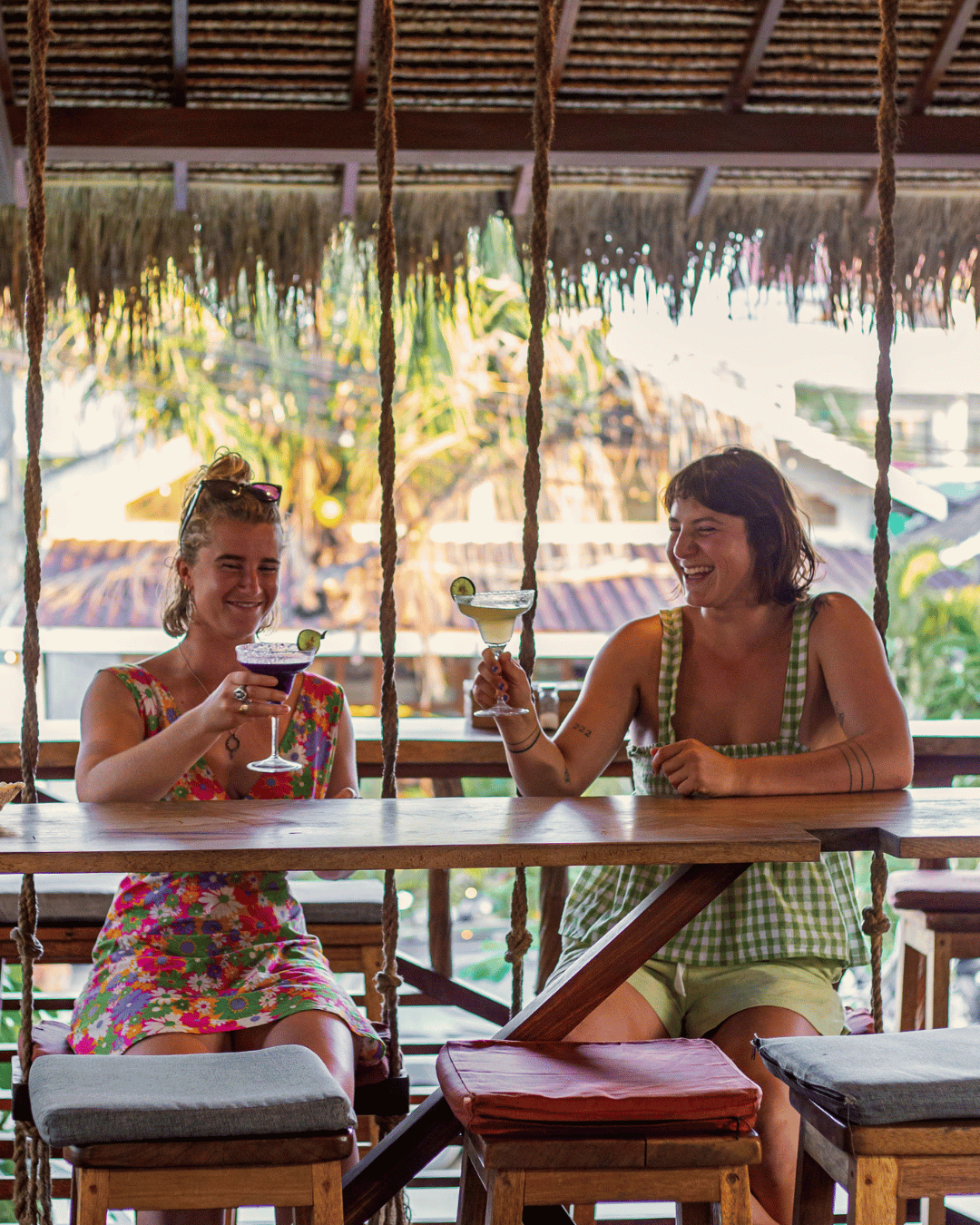 Two women sitting at a wooden bar, enjoying tropical cocktails and smiling. The background shows a thatched roof and lush greenery of Treehouse Restaurant and Bar in Kuta Lombok.