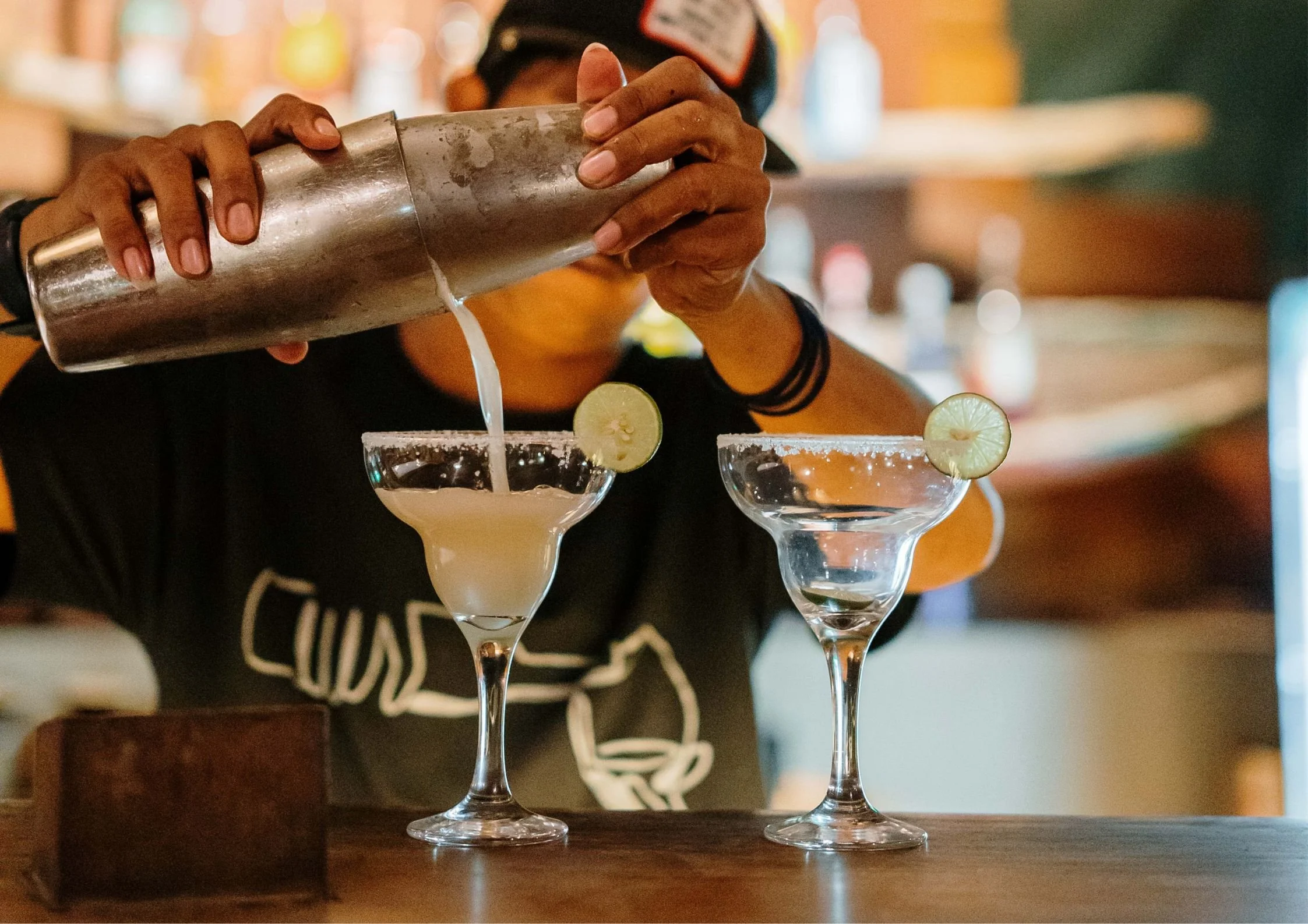 Treehouse bartender pouring a margarita cocktail from a shaker into a glass with a lime wedge, with another similar glass in front.