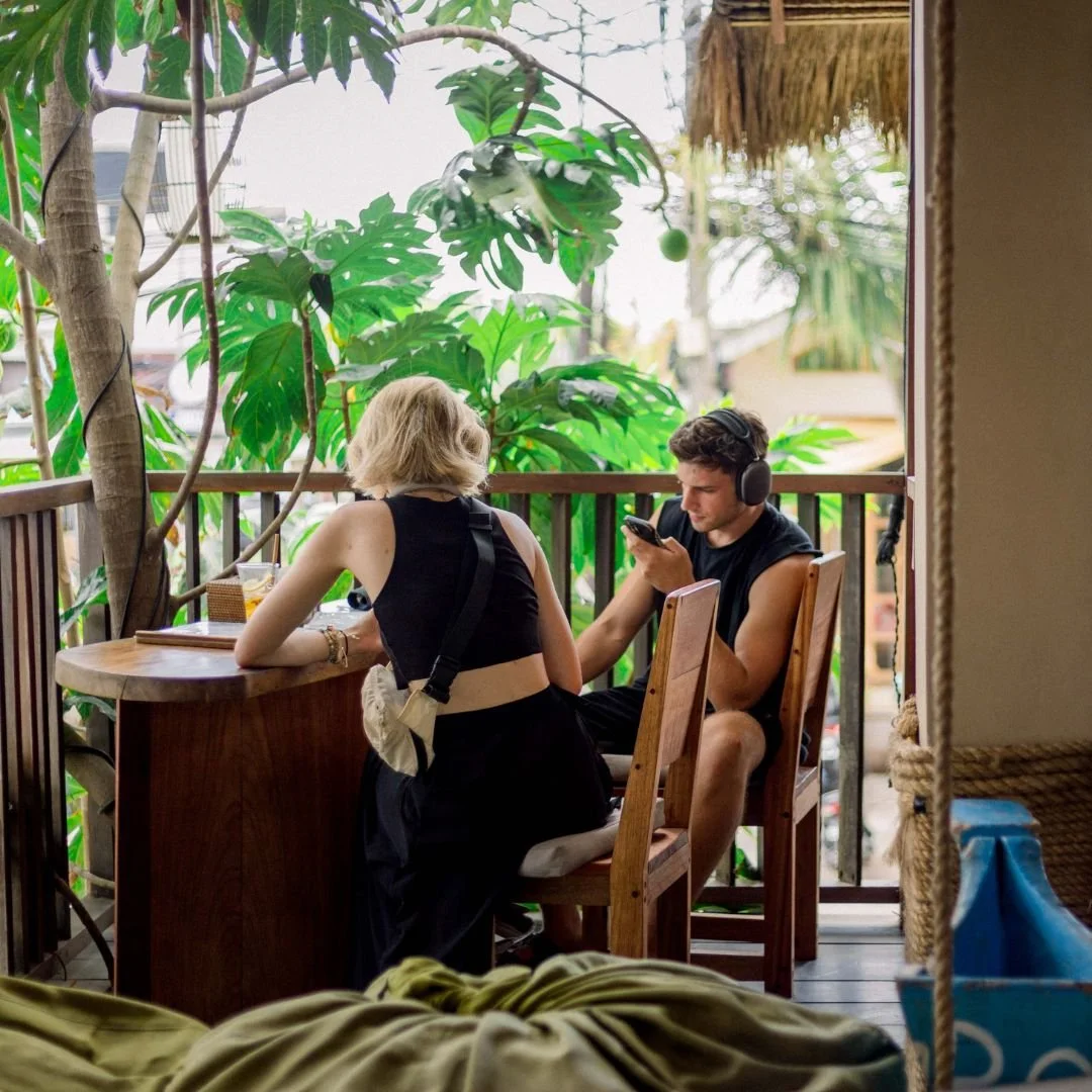 Two young people sitting at a wooden table on a balcony with lush green plants in the background. The woman has blonde hair, wears a black sleeveless top, and has a crossbody bag. The man has dark hair, wears a sleeveless black shirt, headphones, and is looking at his phone. There are drinks on the table, and the setting appears to be a relaxed, outdoor cafe.