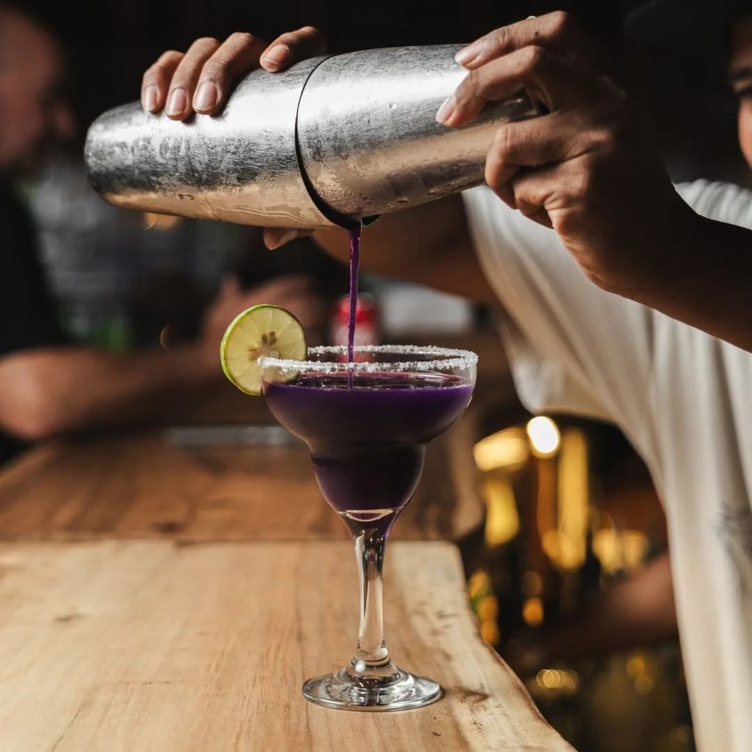 A bartender pours a purple cocktail from a shaker into a glass garnished with a lime wedge and rimmed with salt.