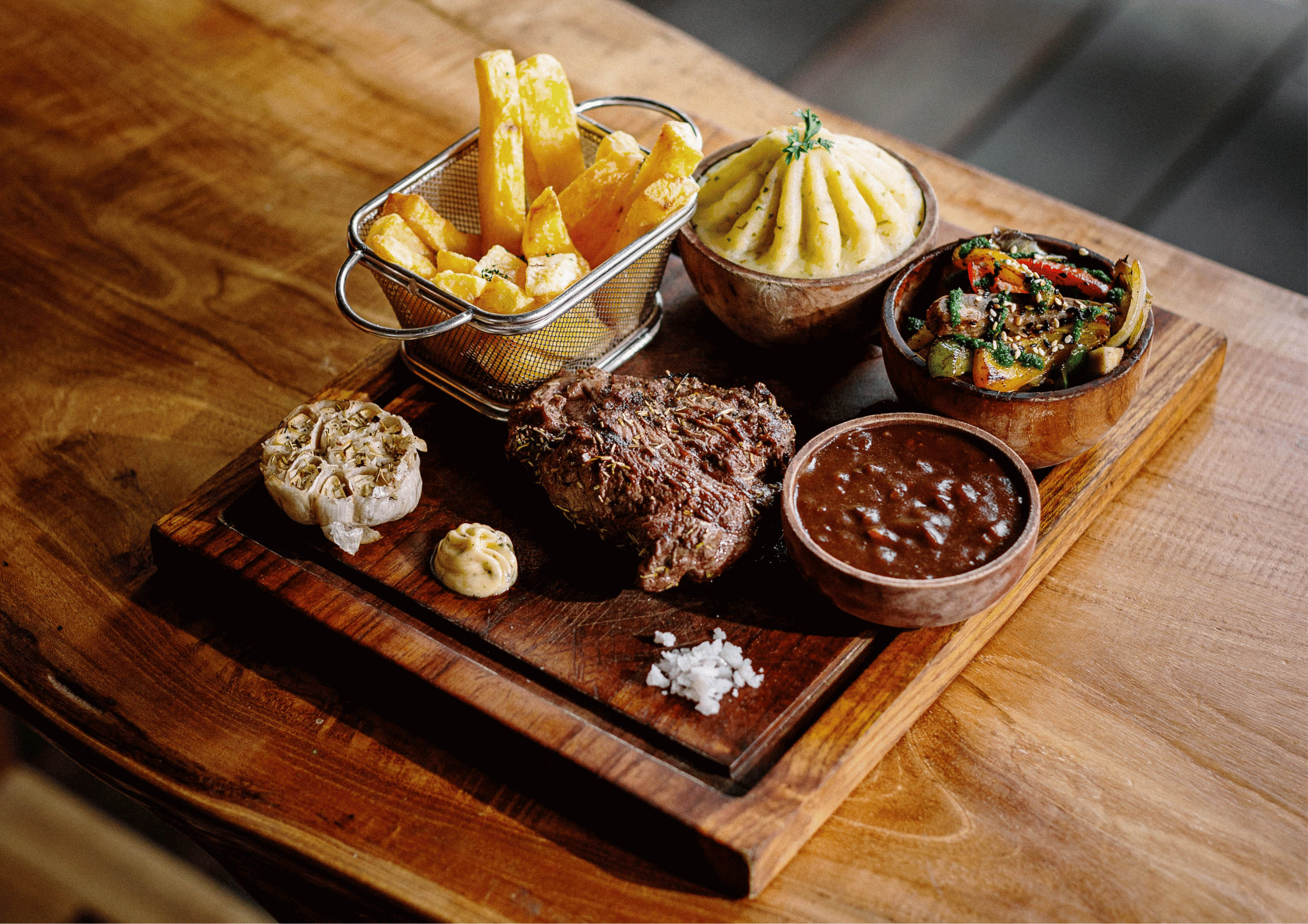 A wooden serving board with tenderloin steak, french fries in a metal basket, mashed potatoes with a small sprig of parsley, and multiple small bowls of gravy and roasted vegetables at Treehouse's Friday Steak Night in Kuta Lombok