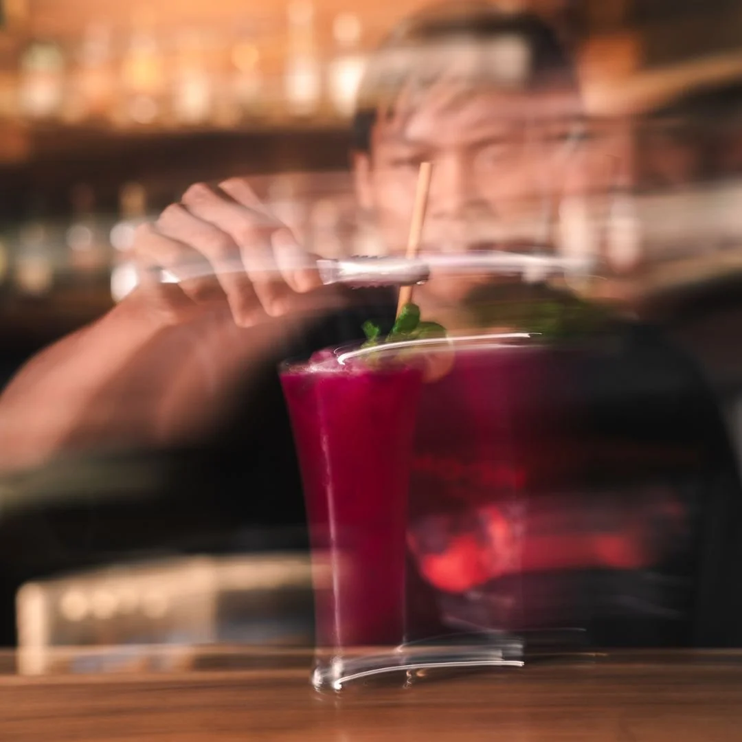 Blurred image of a person drinking a bright red cocktail with a mint garnish at a bar.