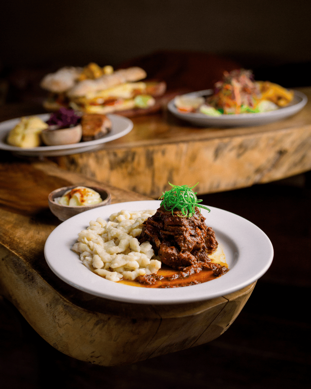 Plate with Hungarian Beef Goulash, dumplings, pickled cucumber salad and chopped green herbs, with additional dishes and appetizers on a rustic wooden table.