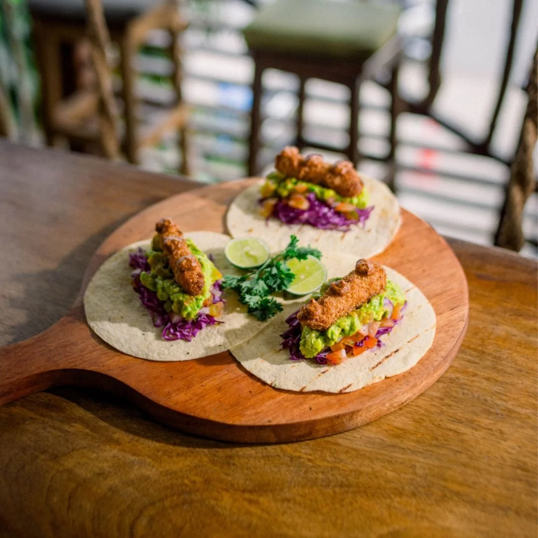Three fish tacos with fried fish, guacamole, purple cabbage, and salsa on corn tortillas, garnished with lime wedges and cilantro, served on a wooden platter.