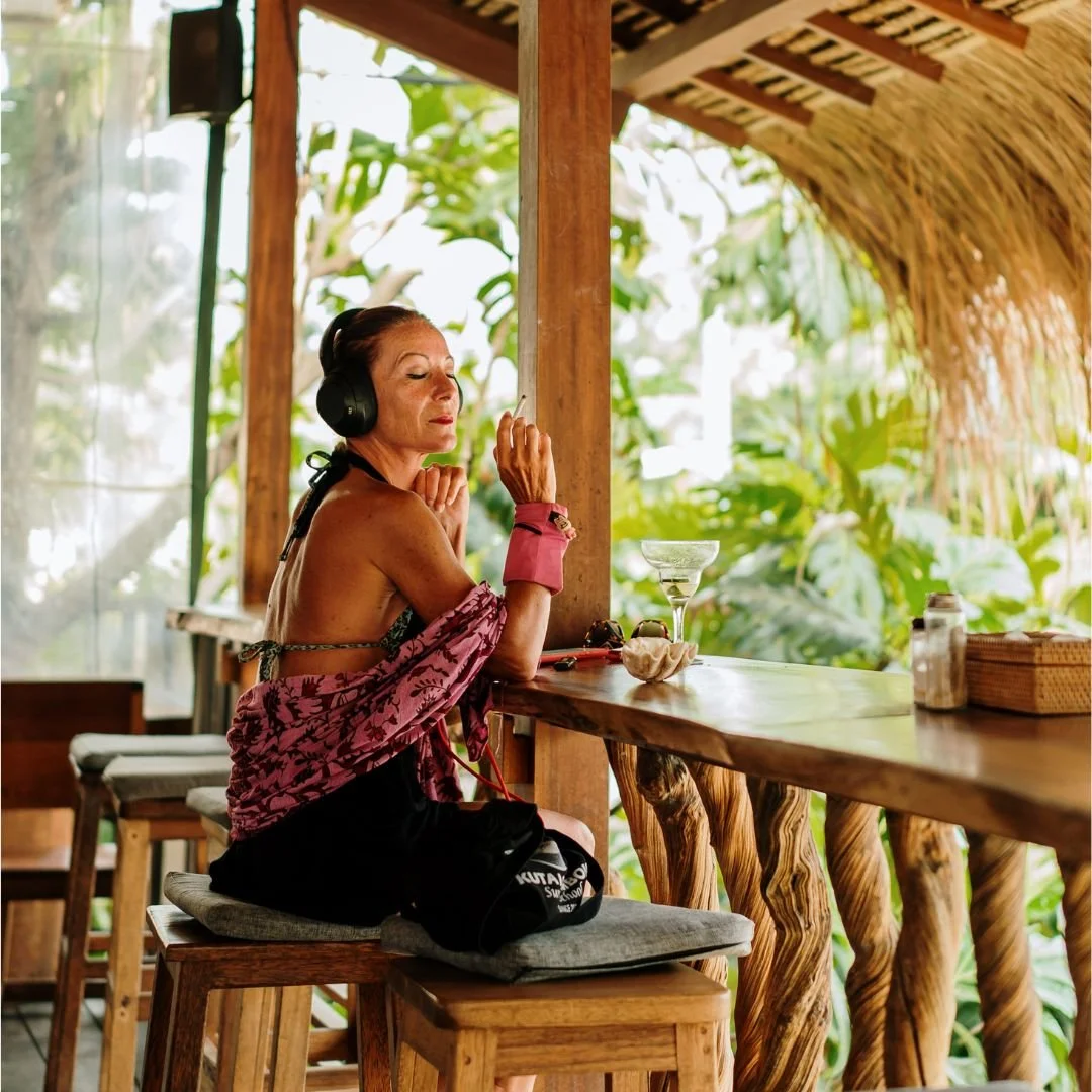An older woman with headphones, seated on a cushion on a wooden stool at a rustic bar, meditating with her eyes closed, in a tropical setting.