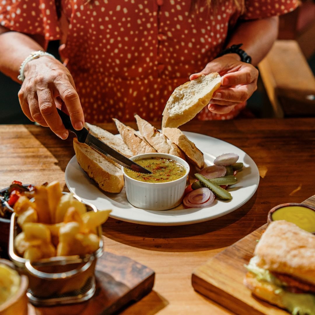 Person spreading bread with butter at a dining table with bread, pickle slices, and a small bowl of sauce or soup.