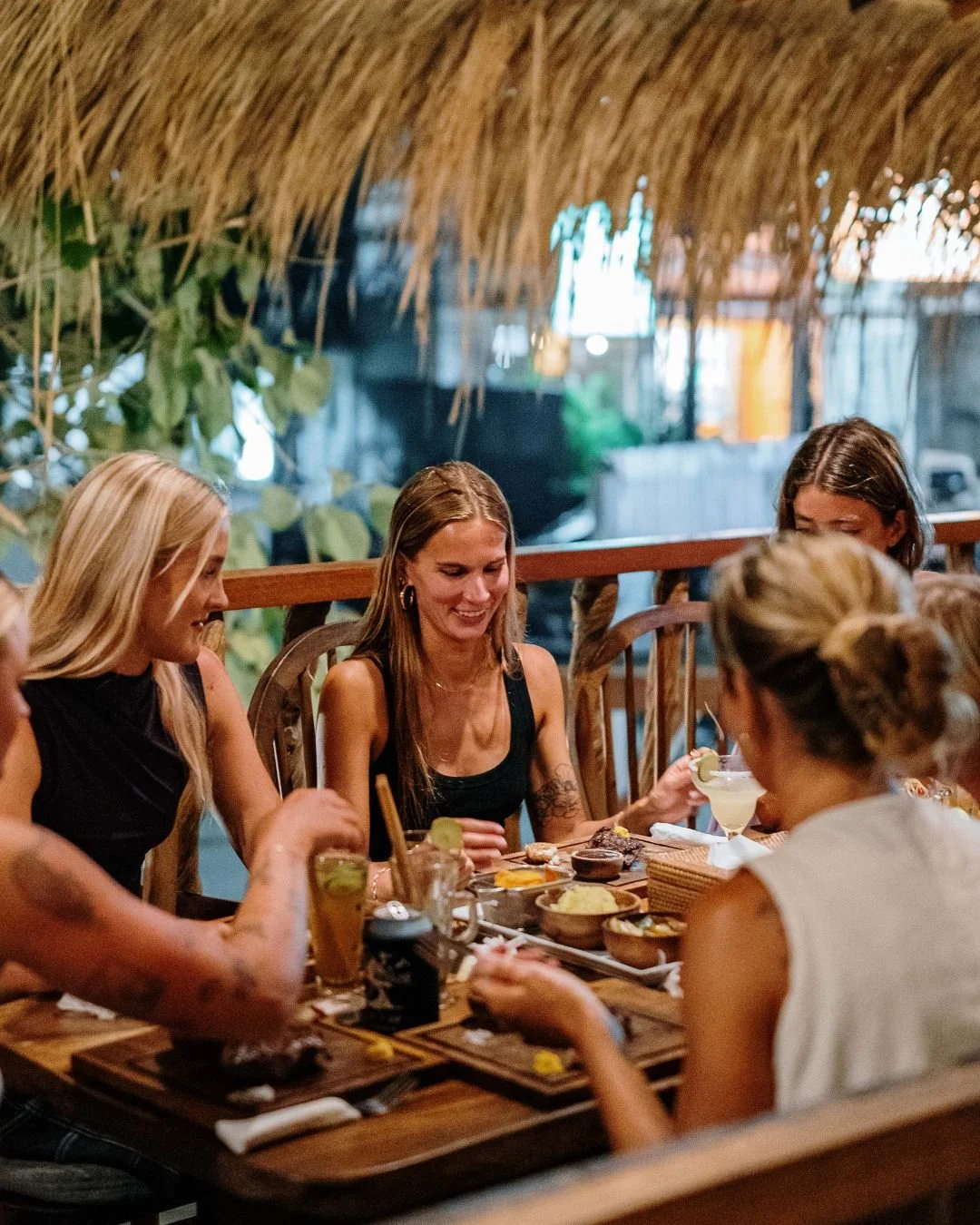 Group of women enjoying a meal at a restaurant with tropical decor, including a thatched roof and lush greenery.