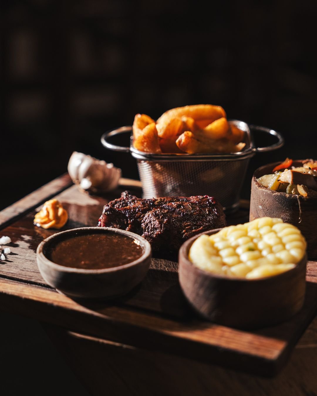 A wooden serving board with assorted foods including a bowl of baked macaroni and cheese, a piece of cooked beef, a bowl of baked beans, fried plantains in a metal strainer, and small bowls of sauce and side dishes.