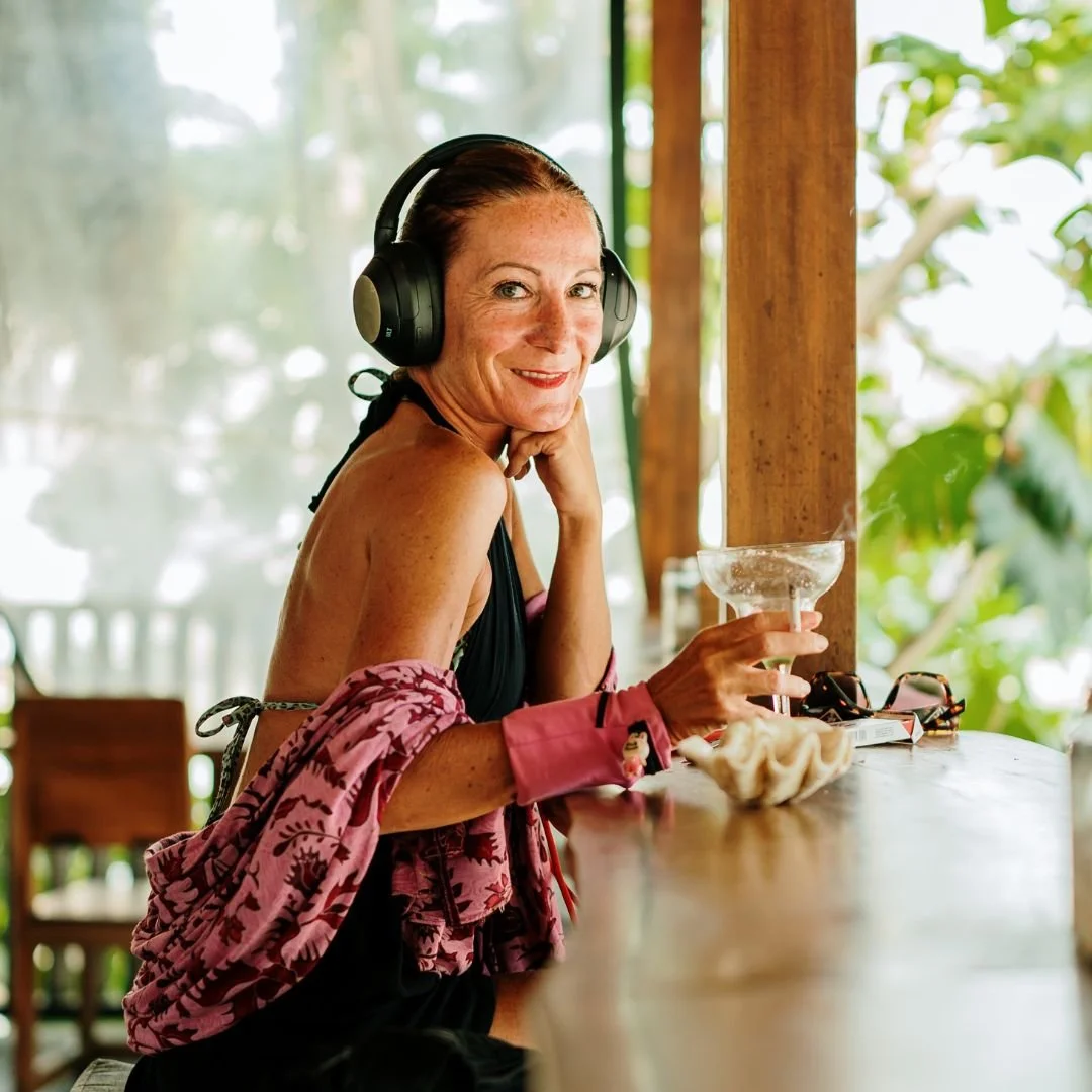 A woman sitting at a wooden bar, wearing headphones and a pink shawl, holding a margarita glass, with a smile, surrounded by lush greenery.