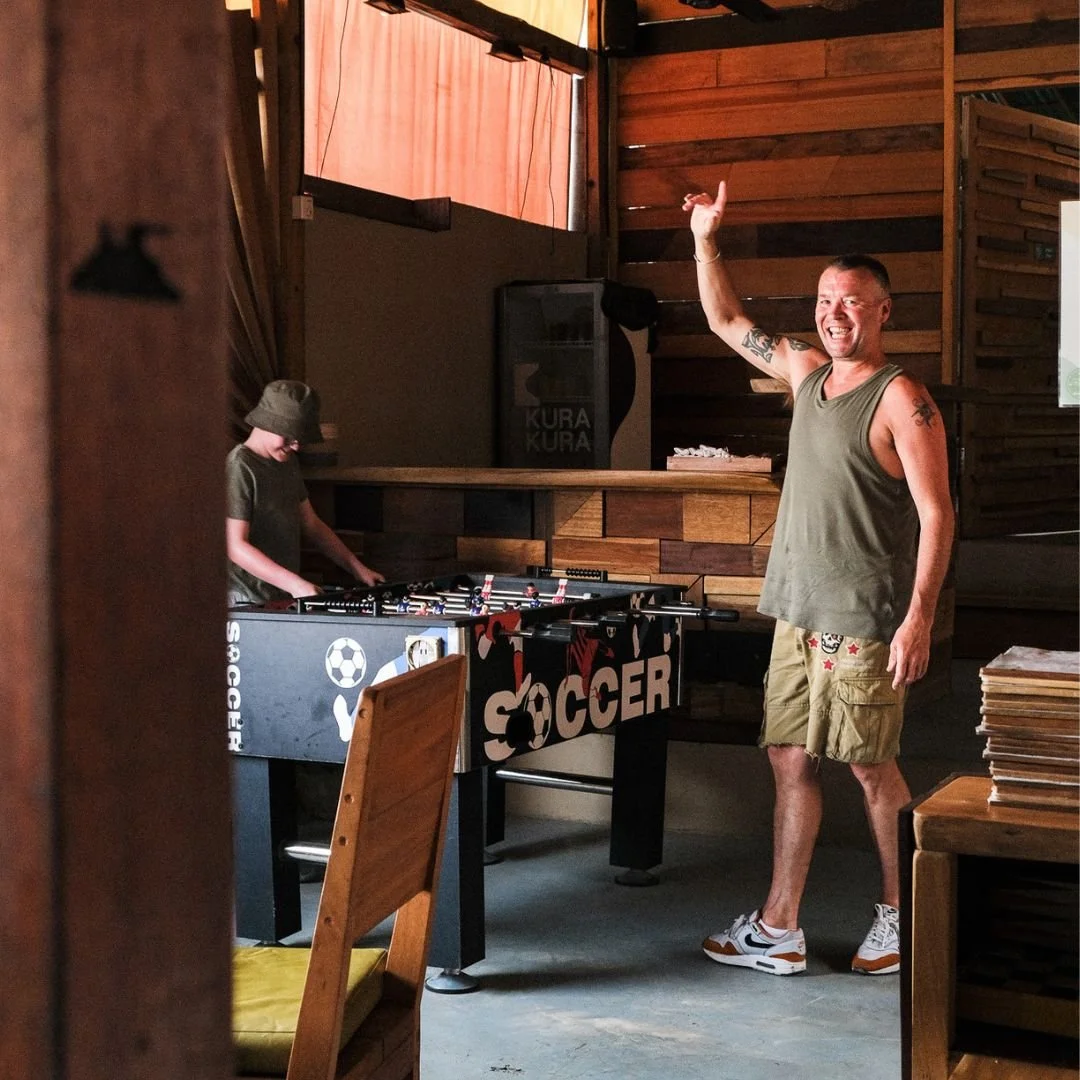 A man with tattoos wearing a green tank top and khaki shorts is smiling and gesturing with one hand while playing foosball with a child in a casual indoor setting with wooden walls.
