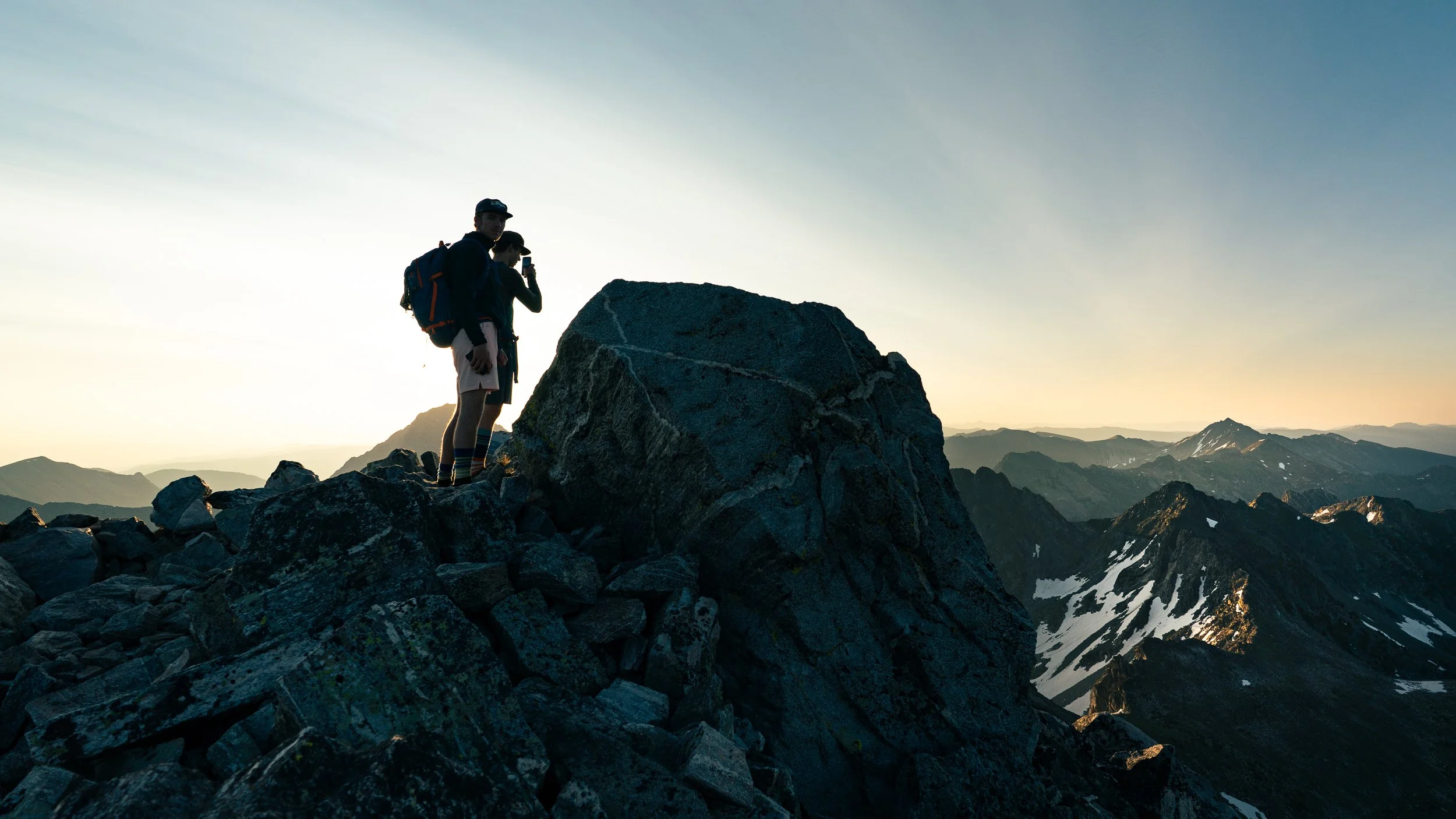 Two hikers standing on rocky mountain terrain at sunset, overlooking distant mountains with snow patches.
