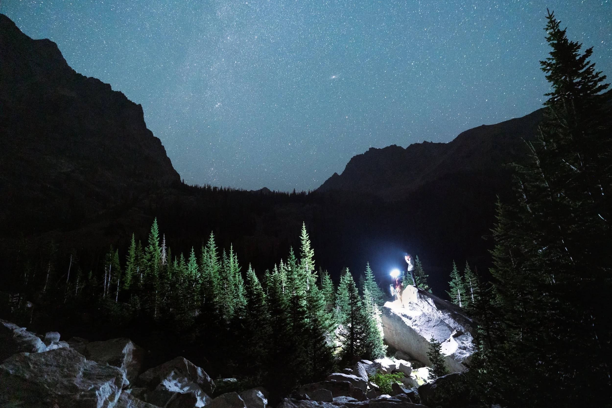 Nighttime mountain landscape with a star-filled sky, dense forest, and a person with a flashlight standing on rocks in the foreground.