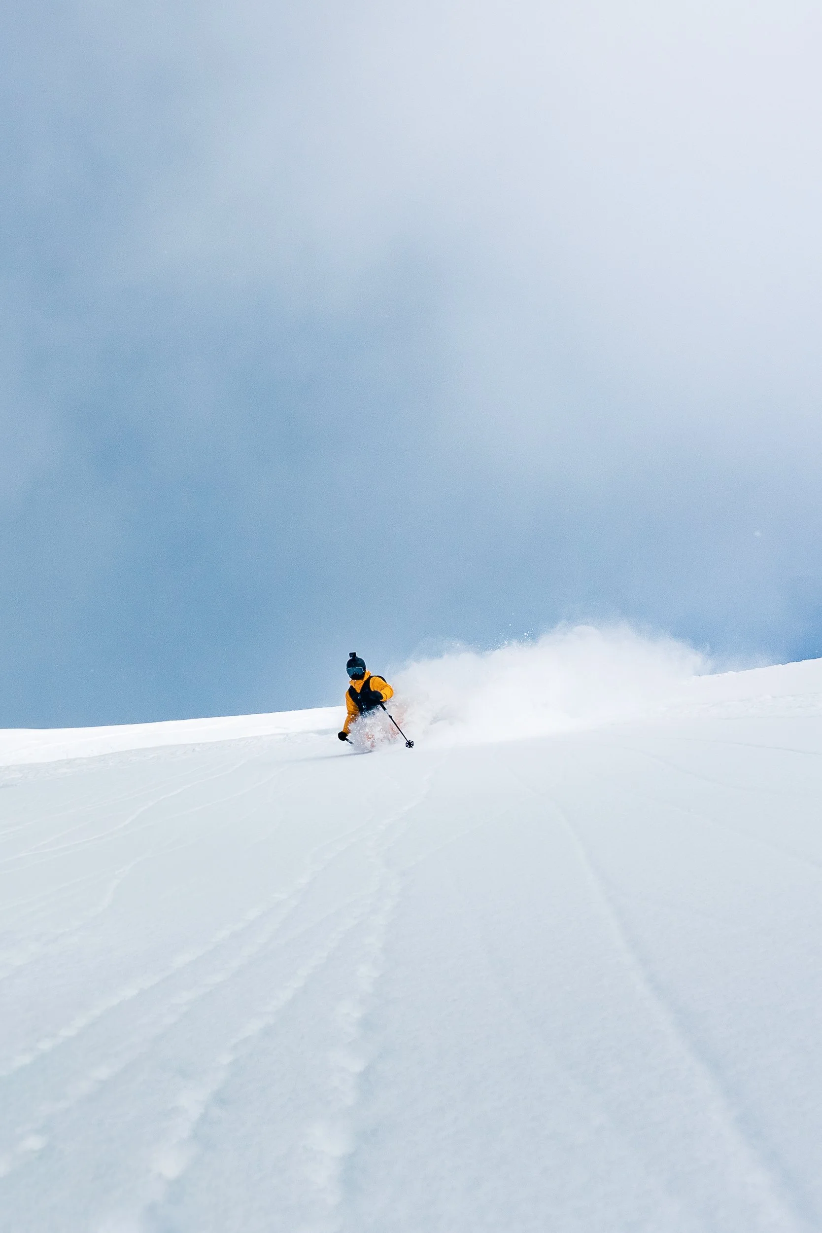 A person in a yellow jacket and blue helmet skiing downhill on a snowy slope under a clear blue sky.