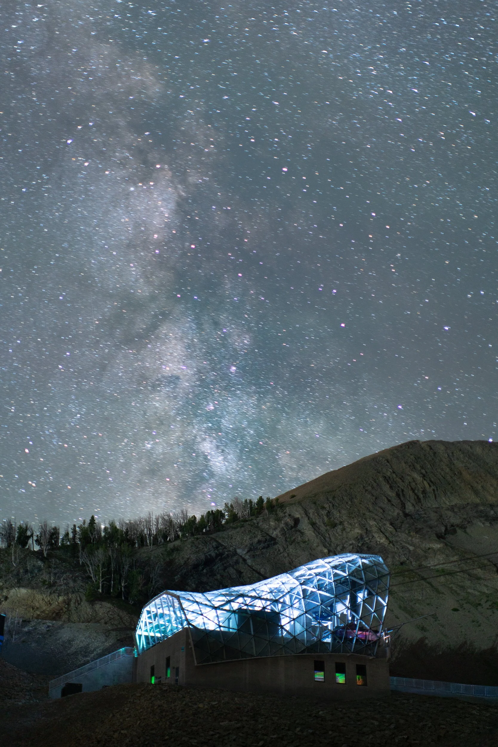 Night sky filled with stars and the Milky Way galaxy above a modern building with geometric design and illuminated windows, located at the base of a hilly landscape with trees.