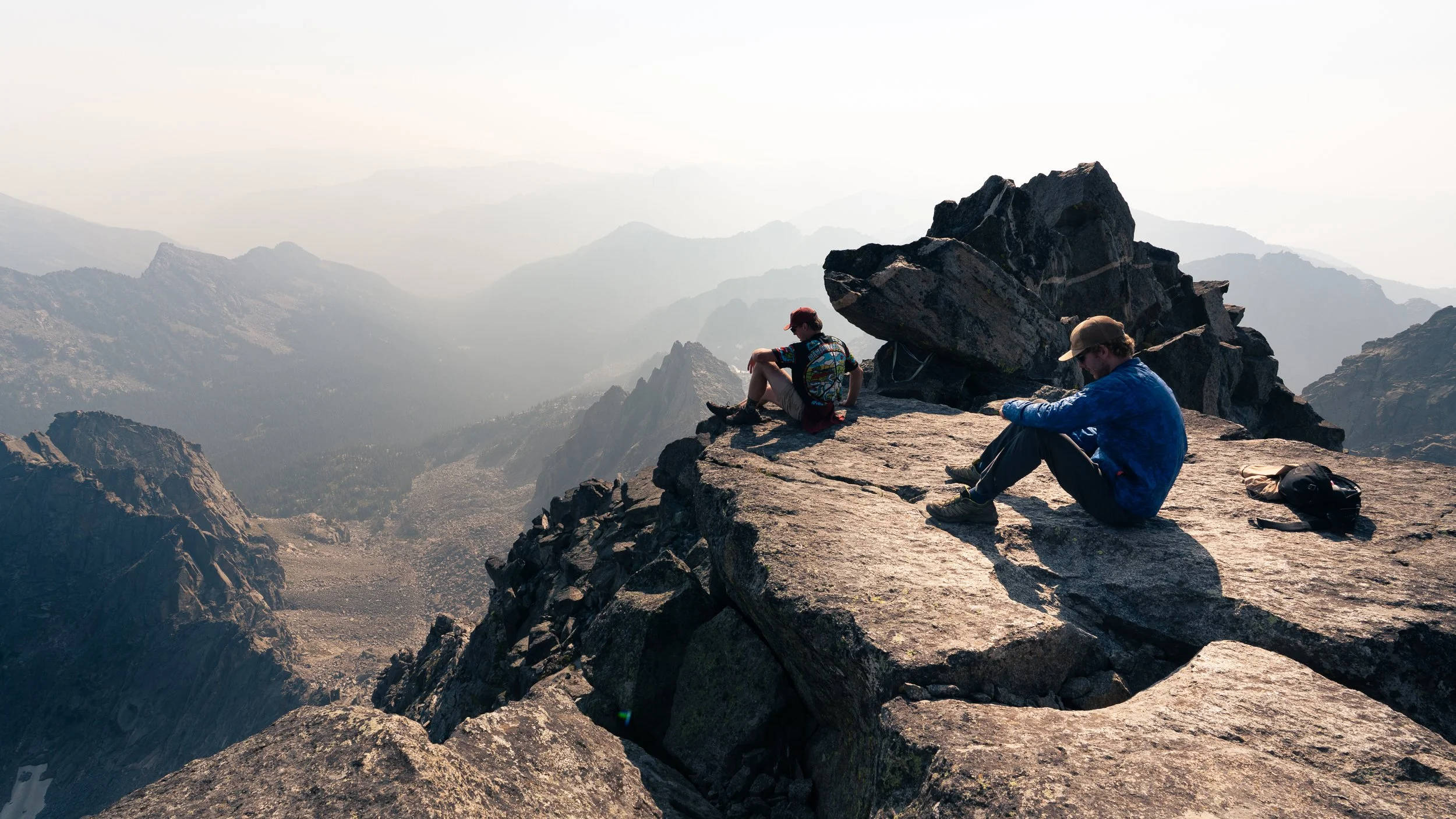 Two hikers sitting on a rocky mountain ledge overlooking a mountain range in the distance.