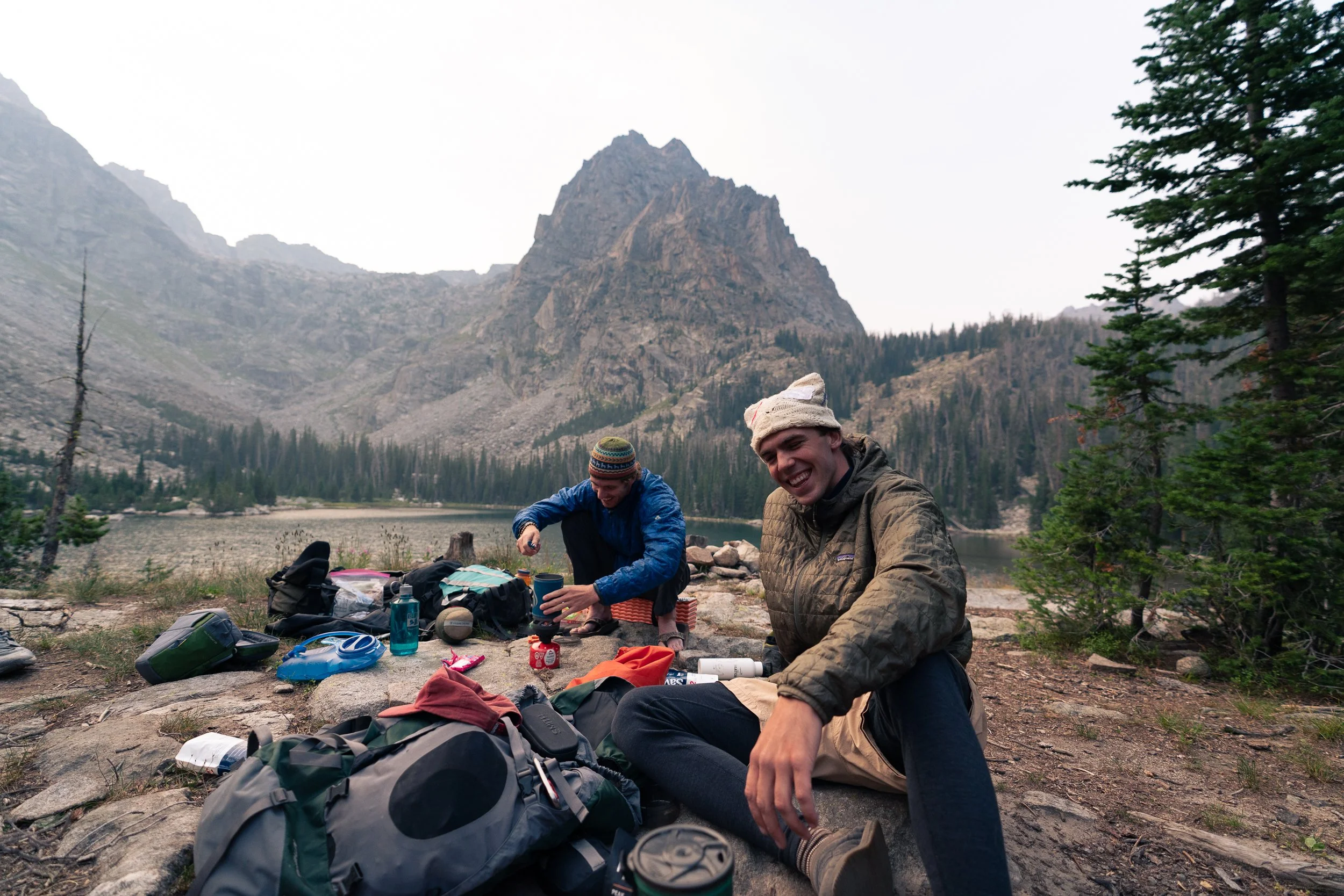 Two men are sitting on the ground near a lake in a mountainous forested area, with gear and supplies around them, smiling and enjoying their outdoor camping trip.