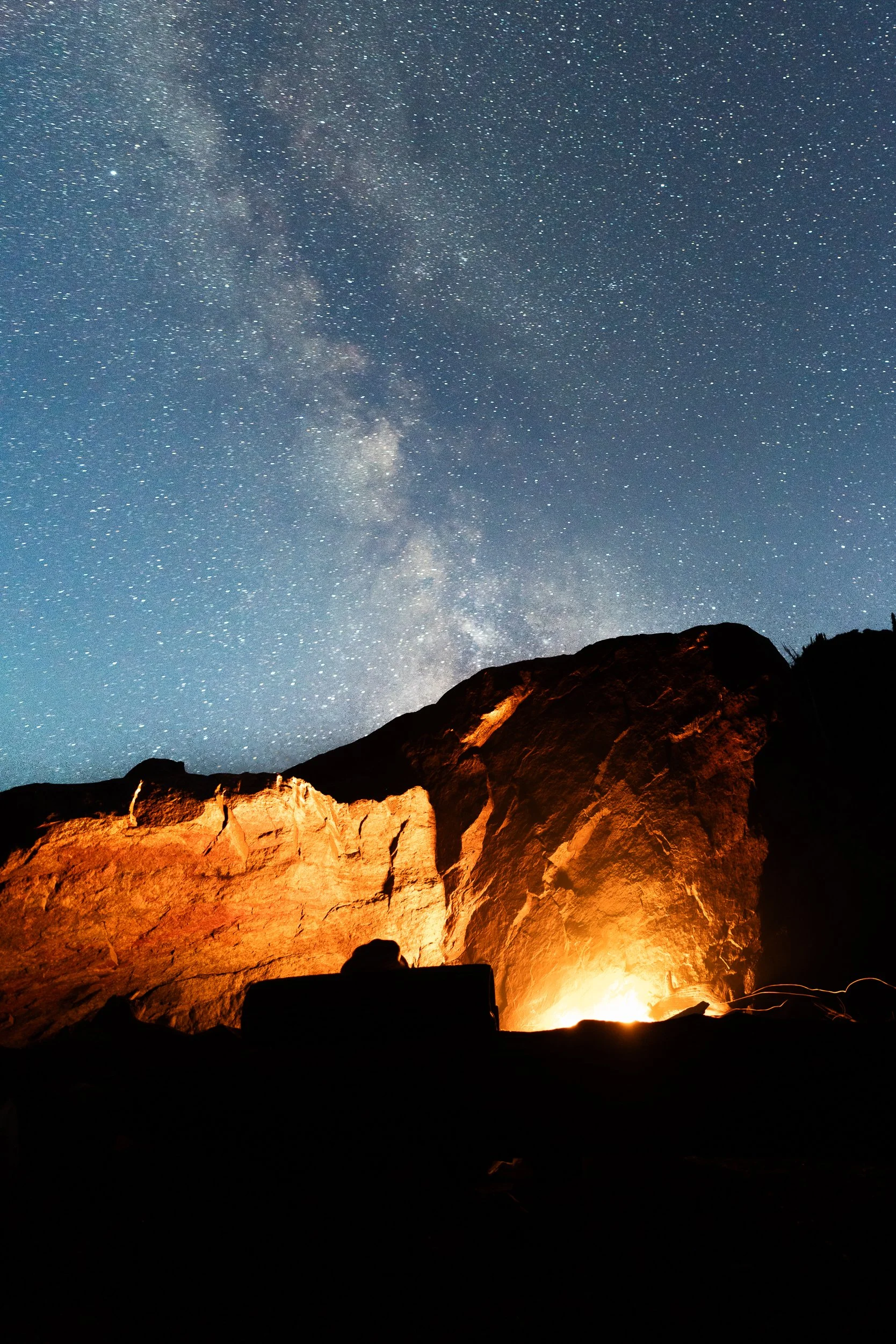 Night sky filled with stars and the Milky Way galaxy above a rocky landscape with a campfire illuminating the rocks.