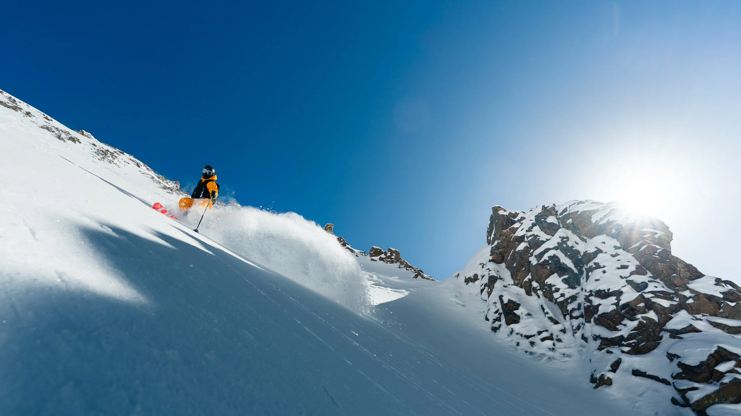 A skier in a yellow jacket and black helmet skiing downhill on snowy mountains under a clear blue sky with the sun shining brightly.