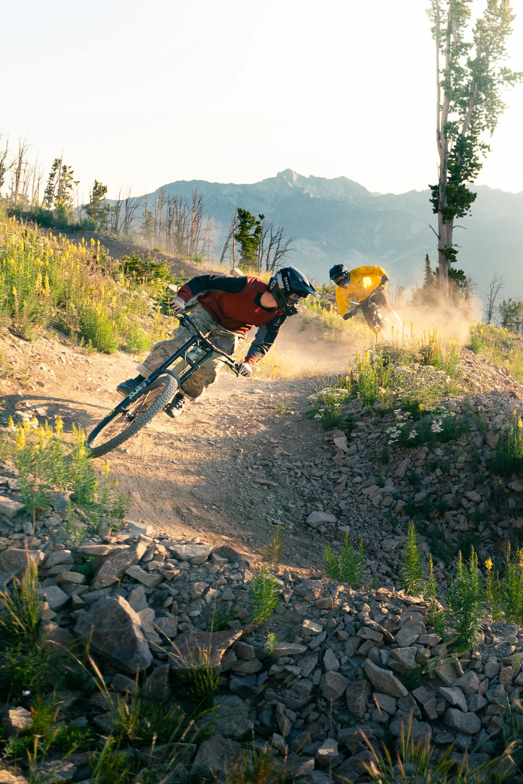 Two mountain bikers on a rocky trail in a mountainous area with tall trees and mountains in the background, dust kicked up from their tires.