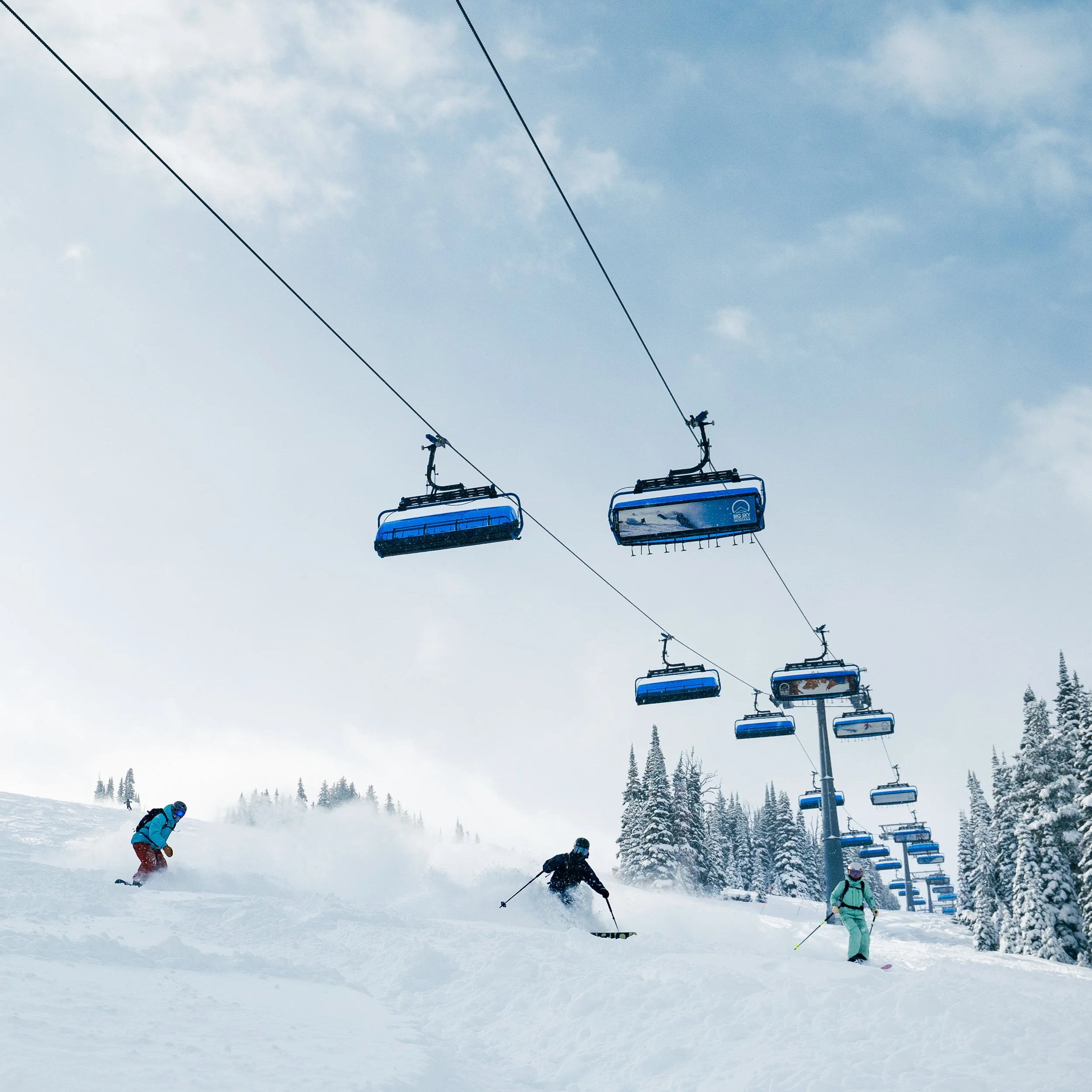 Multiple skiers skiing down a snow-covered mountain near ski lifts on a cloudy day with trees in the background.