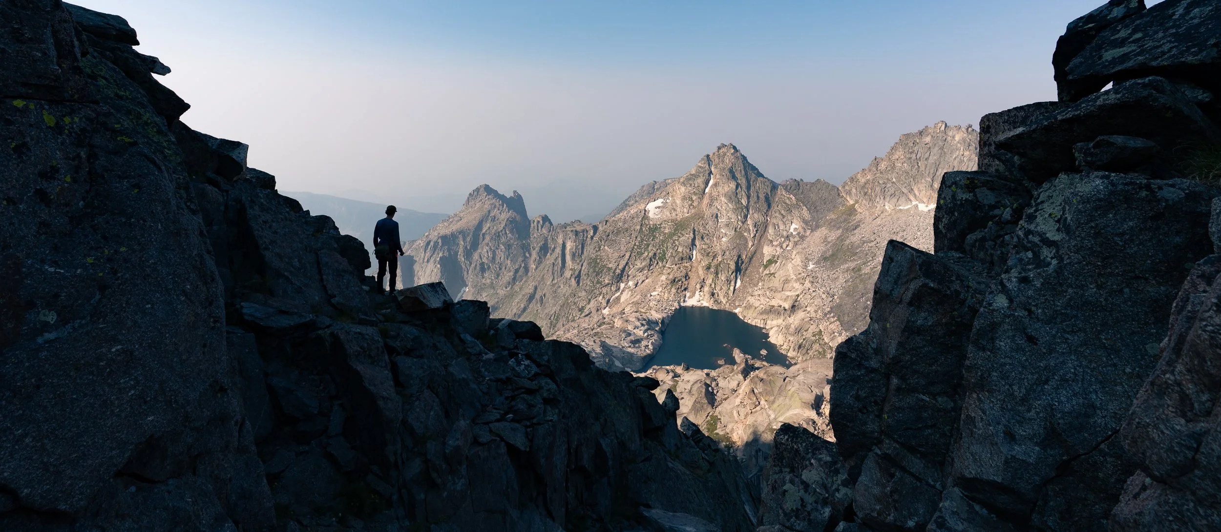 A hiker standing on a rocky mountain trail overlooking rugged mountain peaks and a small mountain lake in a remote alpine landscape.