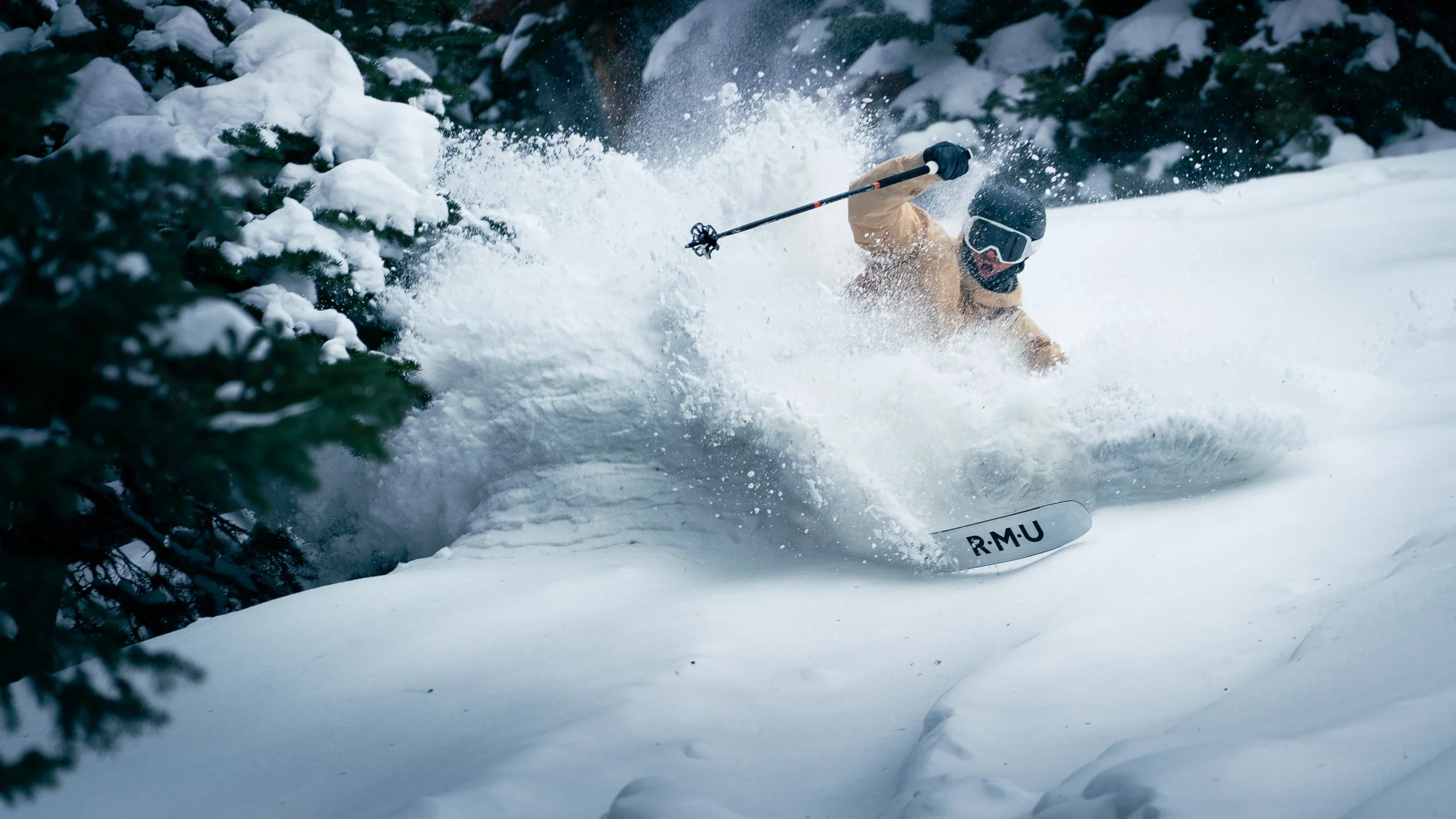 A skier wearing a tan jacket, black helmet, goggles, and gloves skiing down a snowy slope near snow-covered trees.