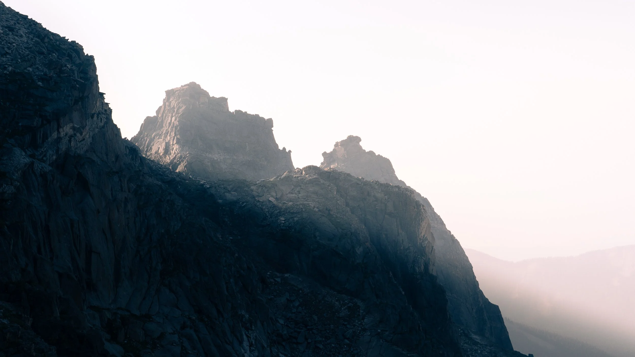 Sunlit rugged mountain range with steep, rocky cliffs and a hazy sky in the background.